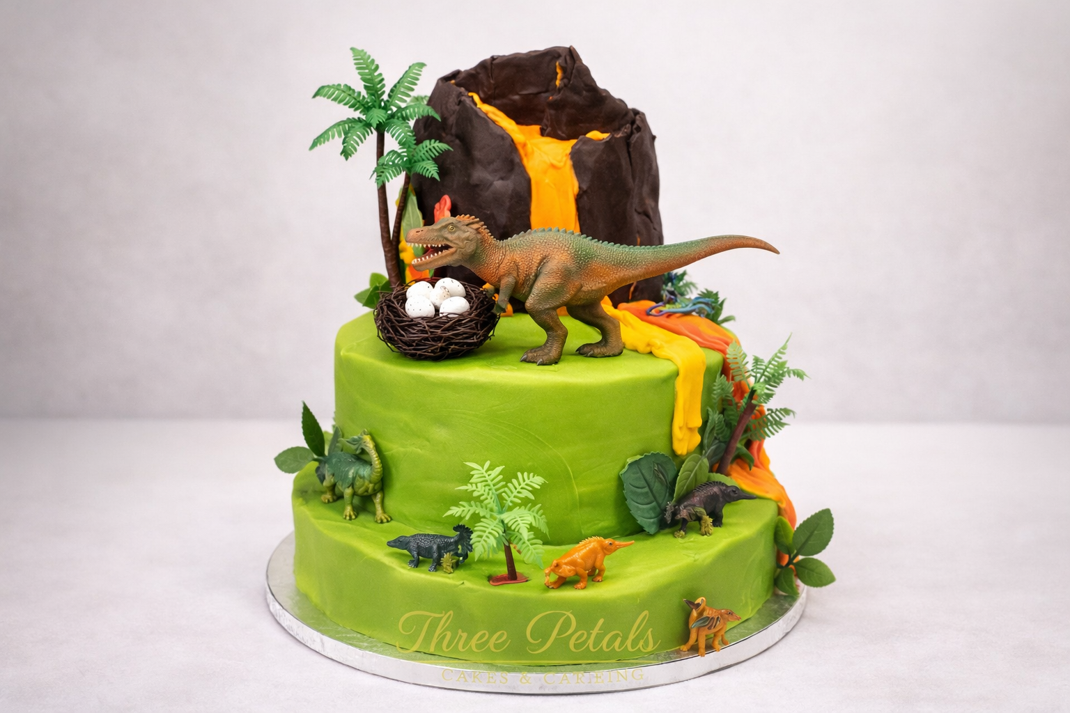 A dinosaur-themed birthday cake with green icing, featuring various toy dinosaurs, palm trees, and a volcano with orange and yellow lava.