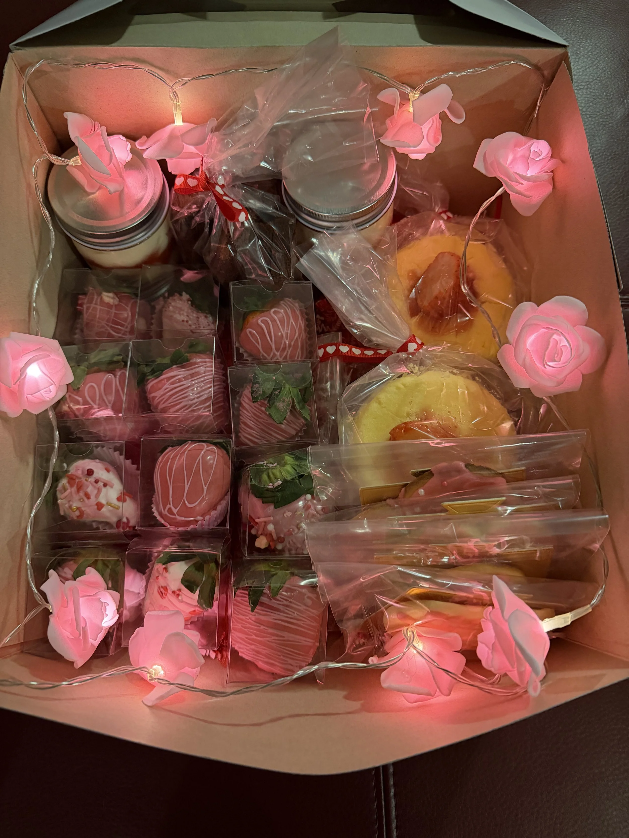 A gift box filled with assorted Valentine's Day-themed chocolates and treats, decorated with pink rose-shaped fairy lights.