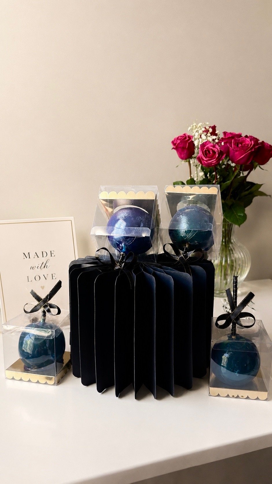 Boxes of blue marbled swirled candles, arranged on a black folded accordion-style display, with pink roses and a 'Made with Love' sign in the background.
