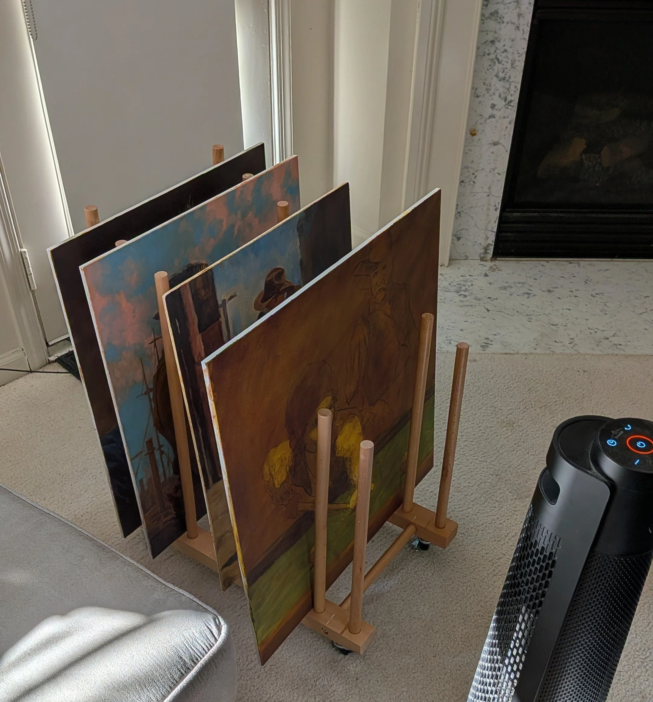 A wooden art display rack holding several paintings, situated near a fireplace and a portable heater on the carpeted floor.