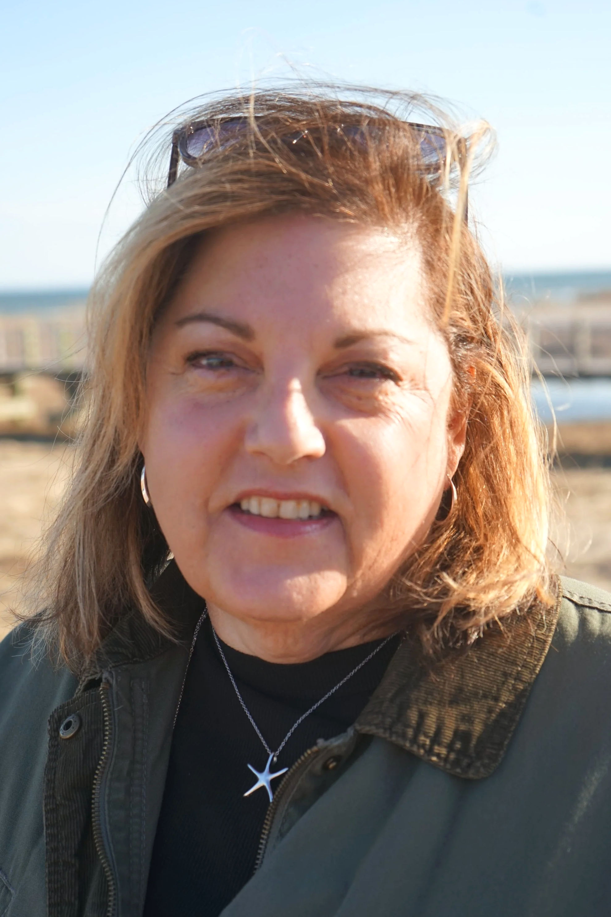 Close-up of a middle-aged woman with shoulder-length hair outdoors on a sunny day, wearing sunglasses on her head and a necklace with a star-shaped pendant.