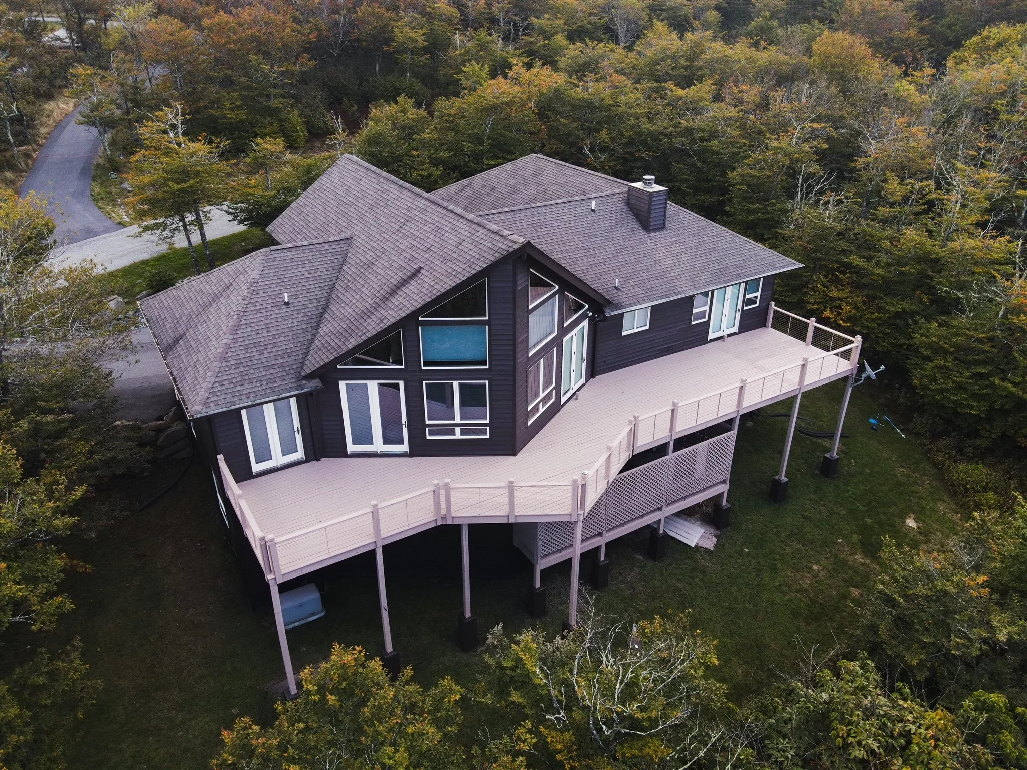 An aerial view of a modern black house with large windows and a spacious wooden deck, situated among trees with autumn-colored leaves.