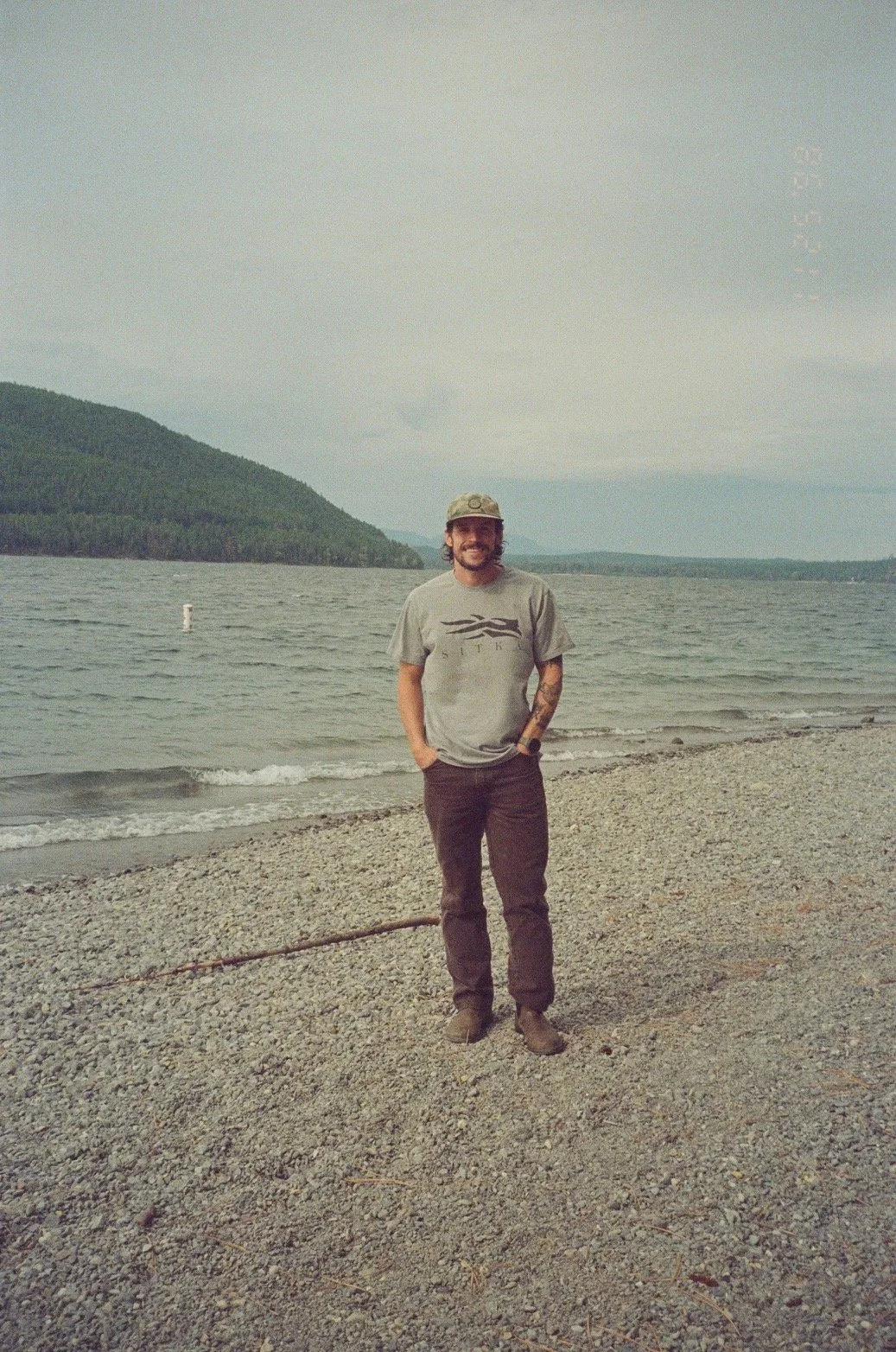 A man standing on a pebbled beach near a lake with mountains in the background, wearing a gray t-shirt, dark pants, and a cap, smiling at the camera.