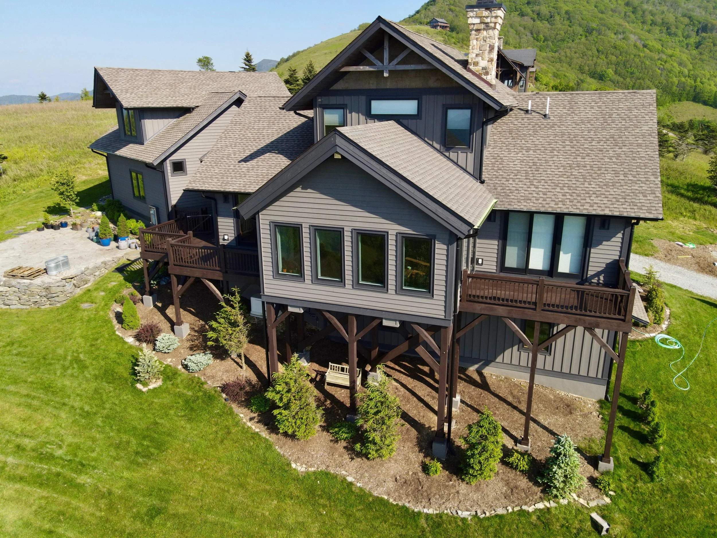 An aerial view of a large, multi-story house with gray siding and a brown roof, situated on a lush green lawn with trees and hills in the background.