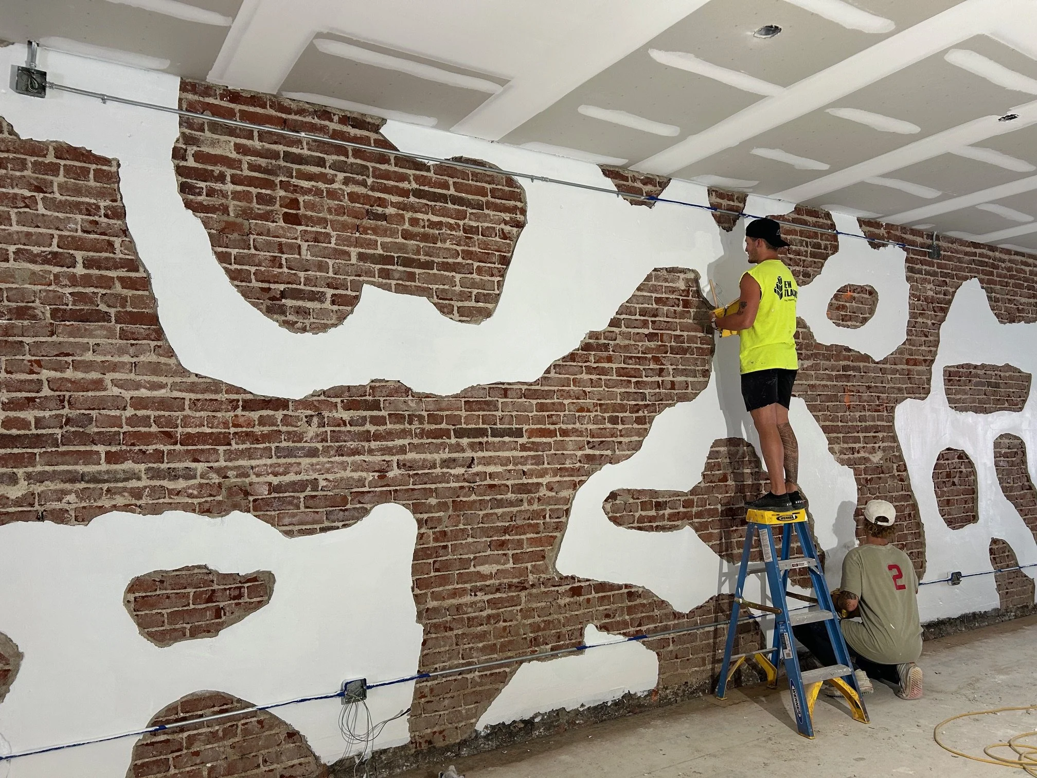 Two workers are installing a decorative wall with exposed red bricks and white paint patches.