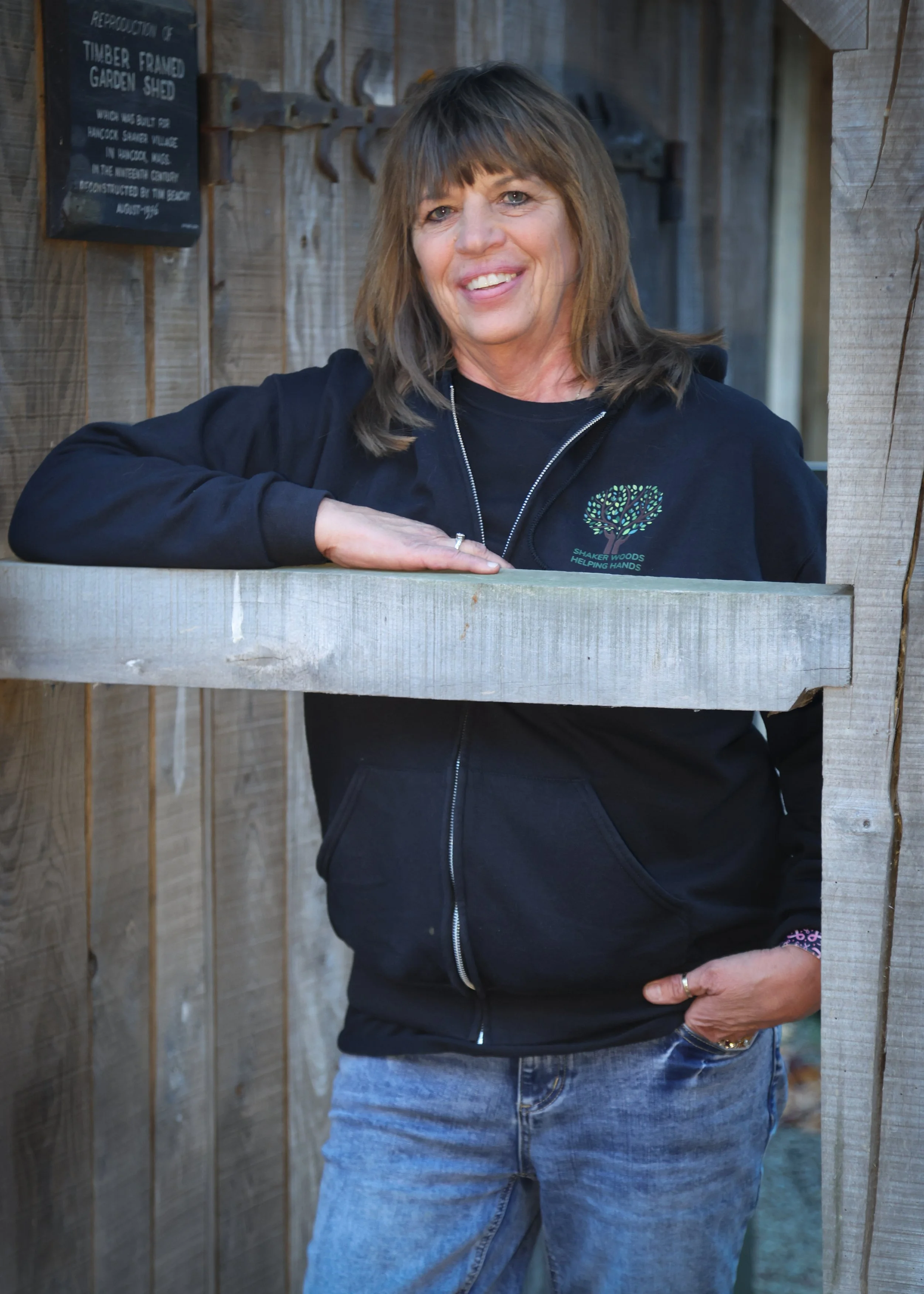 A woman with light brown hair and a black hoodie stands outdoors, leaning on a wooden railing, smiling.