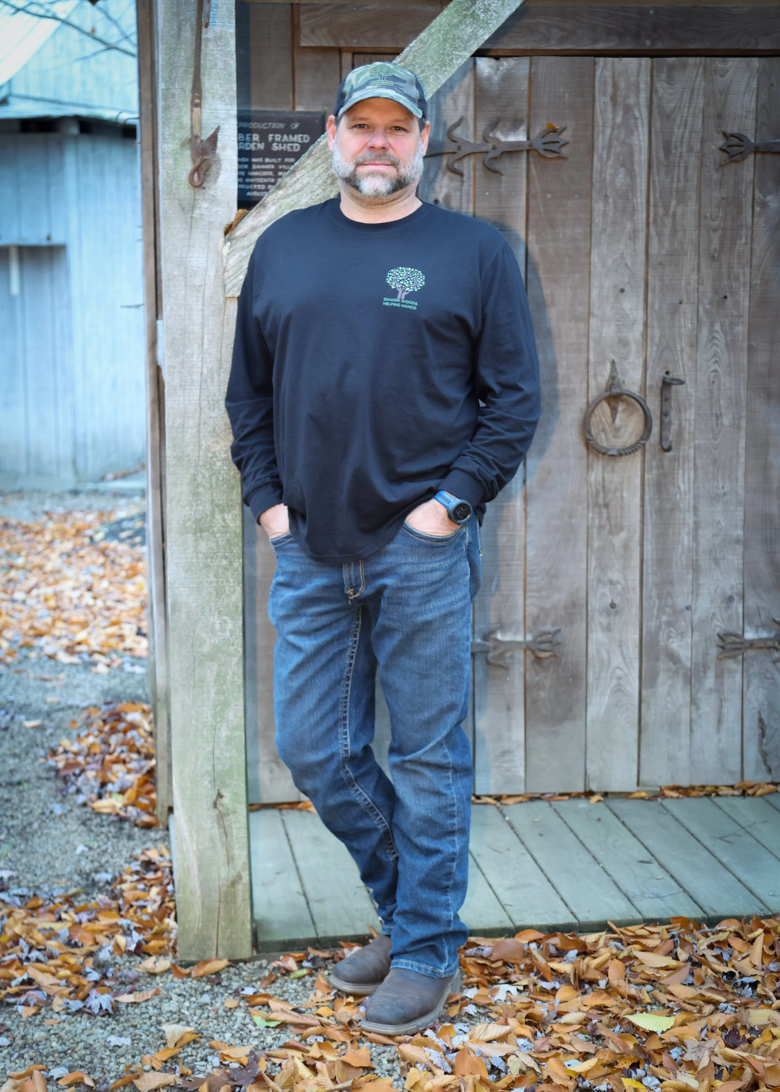 A man with a beard and mustache wearing a camouflage cap, black long-sleeve shirt, blue jeans, gray shoes, and a smartwatch, standing outdoors in front of a wooden shed, surrounded by fallen autumn leaves.