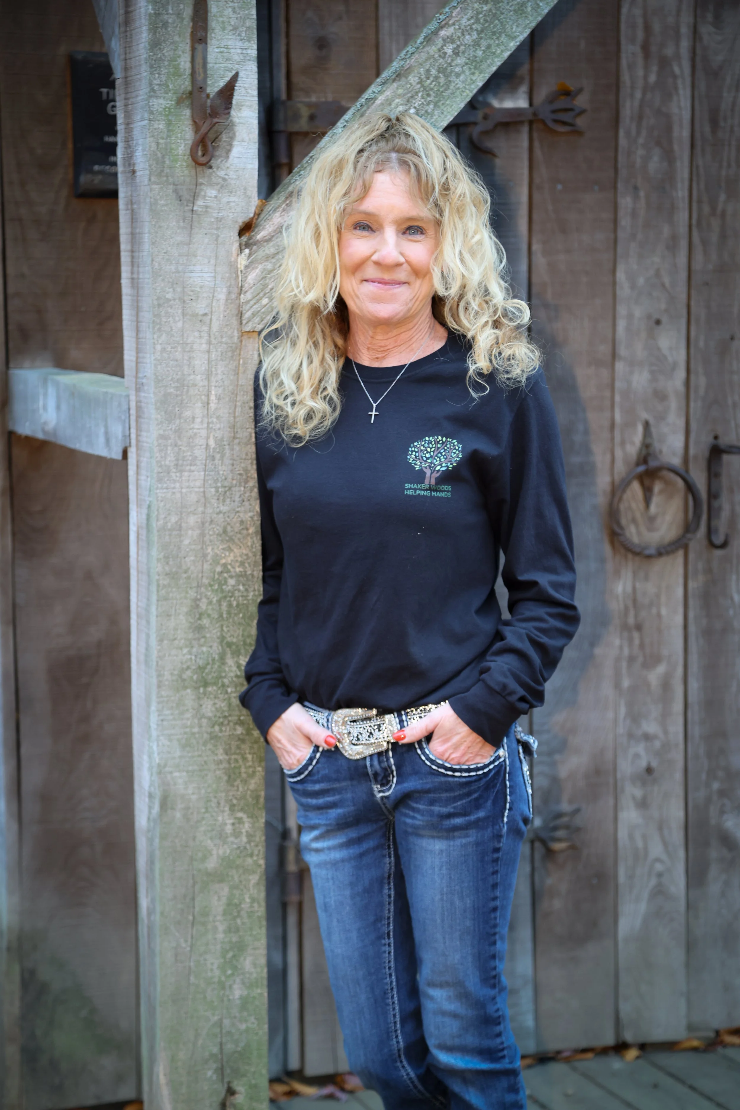 A woman with curly blonde hair wearing a black long sleeve shirt with a logo, jeans, and a decorative belt, leaning against a wooden post in front of a wooden wall.