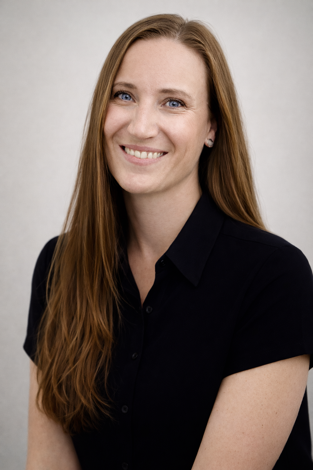 A woman with long red hair, blue eyes, and fair skin, smiling, wearing a black collared shirt, against a plain light gray background.