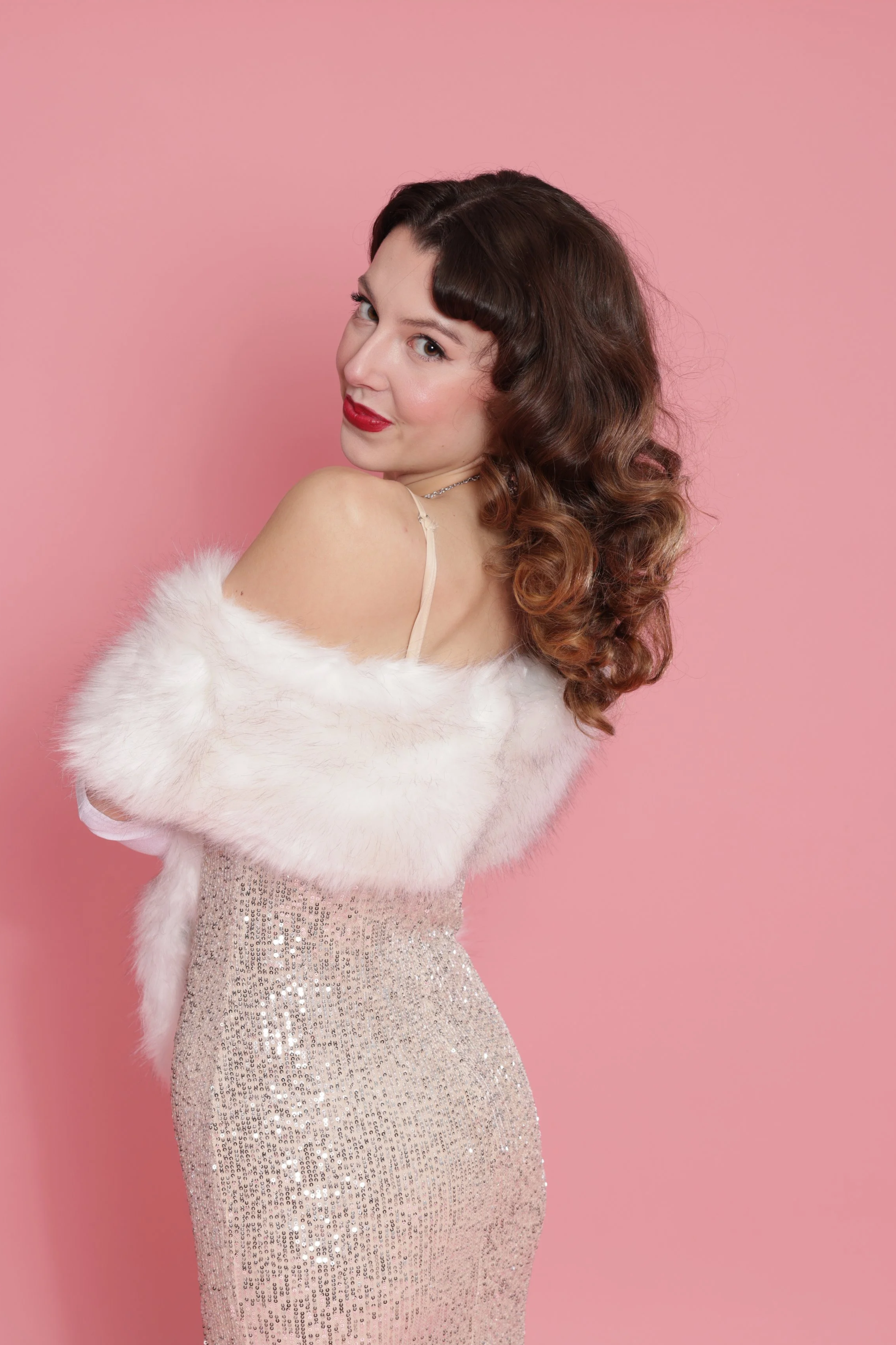 Woman in a sparkly dress and white fur stole, with styled brown hair and red lipstick, posing against a pink background.