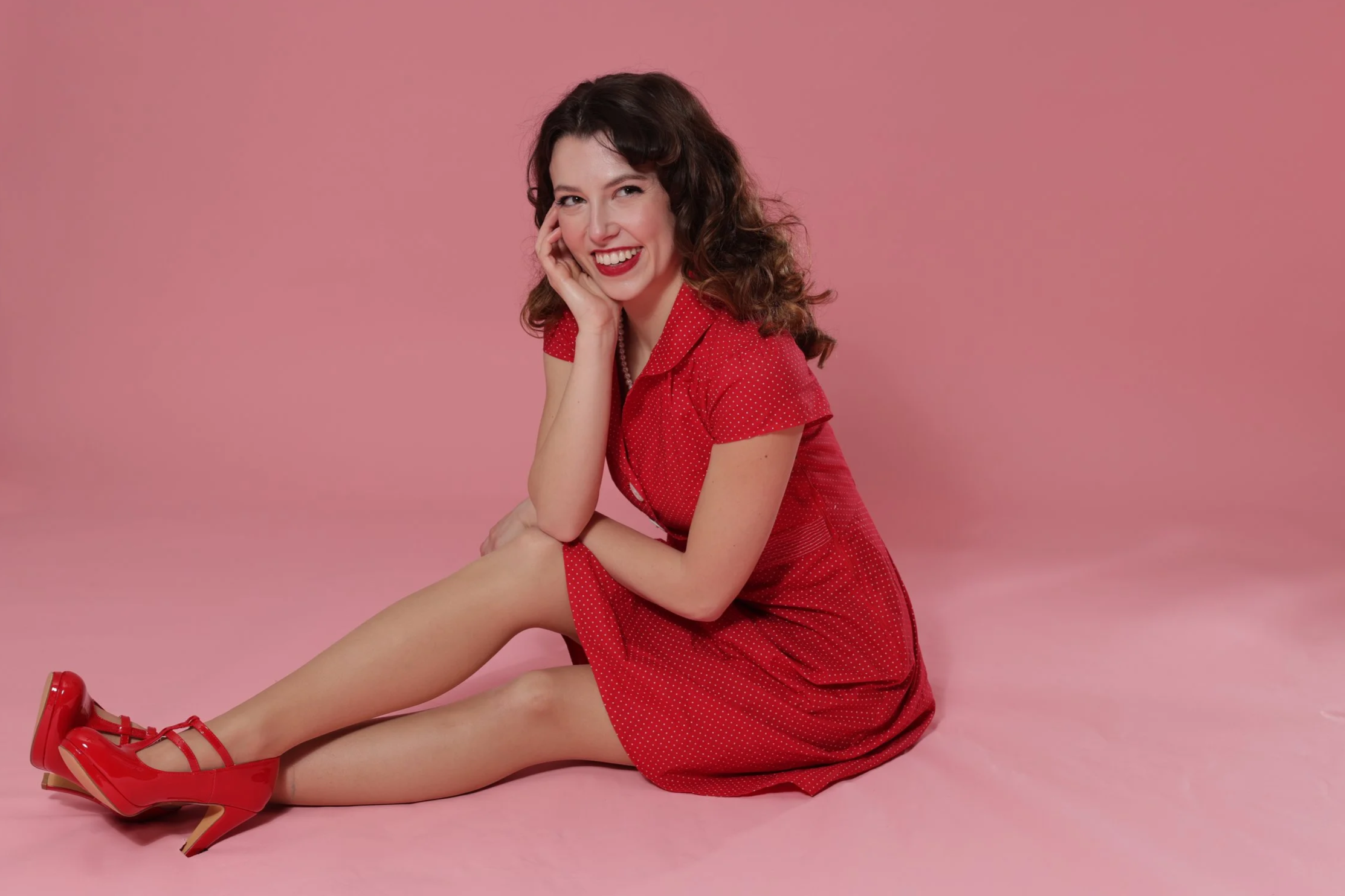 A woman with curly brown hair sitting on the pink floor, wearing a red polka dot dress and red high heels, smiling and winking at the camera.