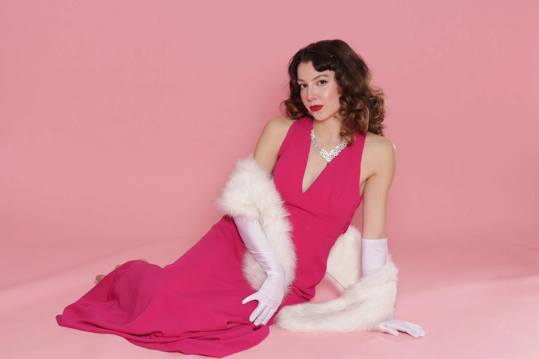 A woman in a pink evening gown sitting on the floor against a pink background, wearing white gloves and a white fur stole, with a silver necklace and red lipstick.
