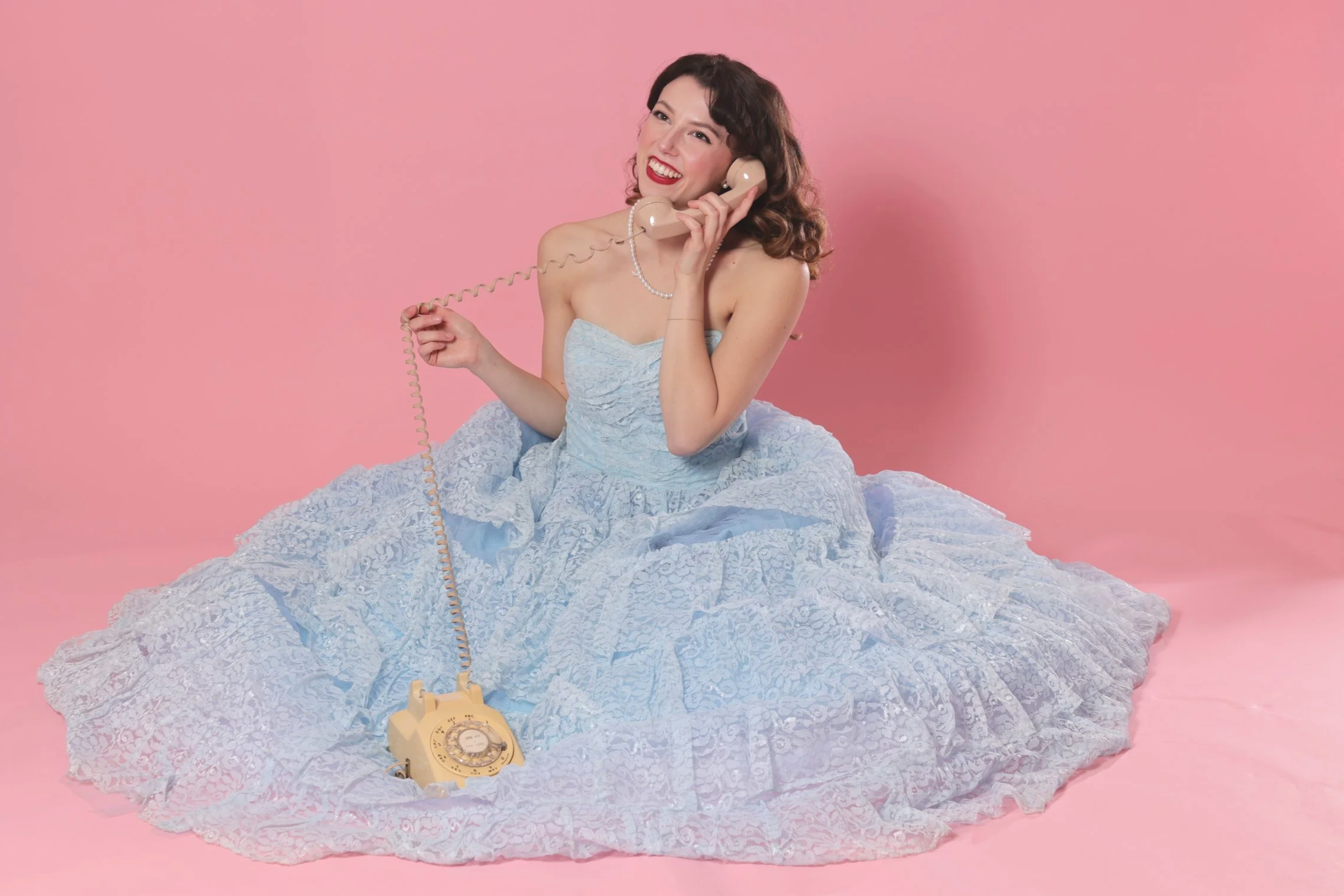 A woman in a light blue lace dress sitting on a pink floor and pink background, using a vintage beige rotary phone, smiling and holding the receiver to her ear.