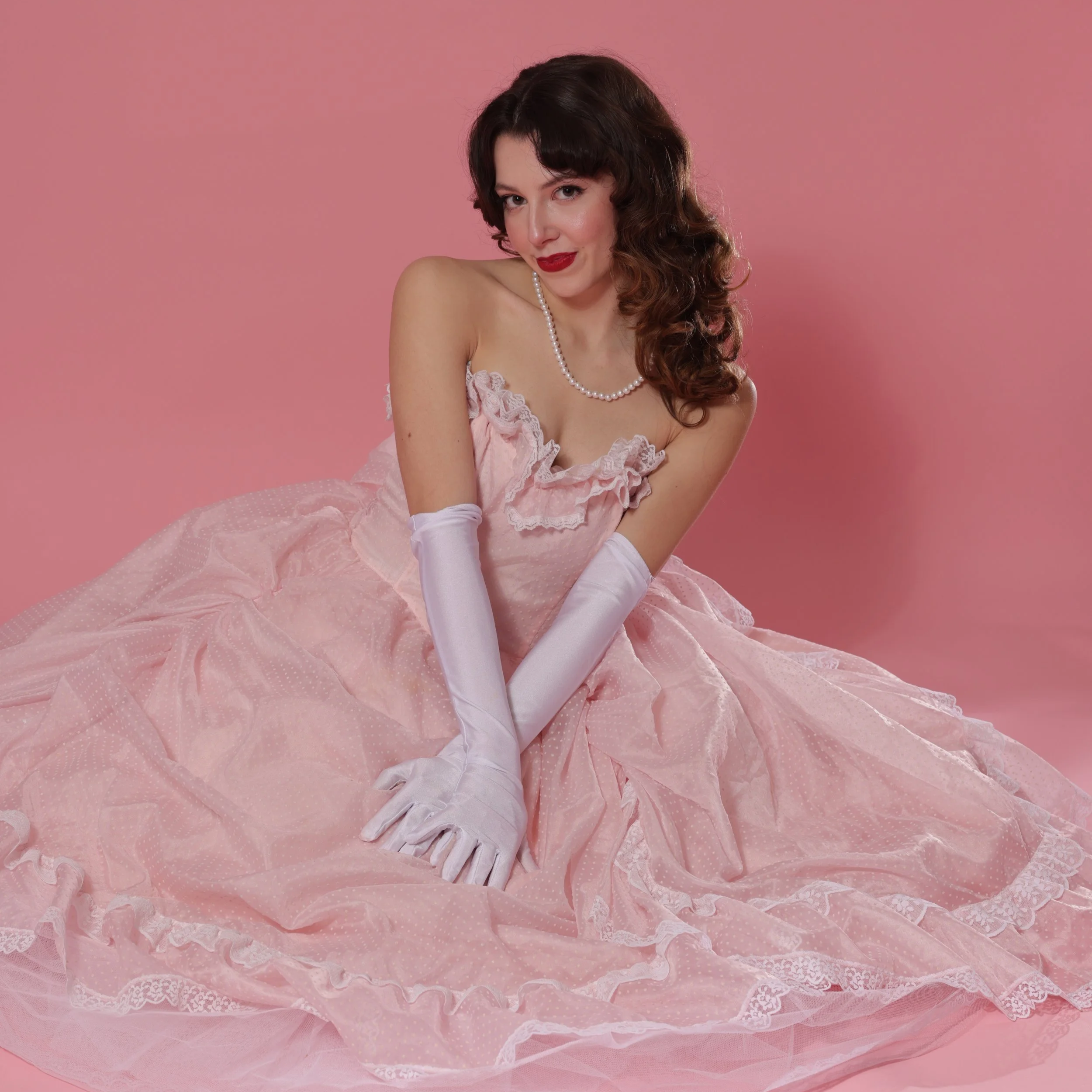 Woman in a vintage pink dress with ruffles and lace, white gloves, pearl necklace, red lipstick, posing against a pink background.