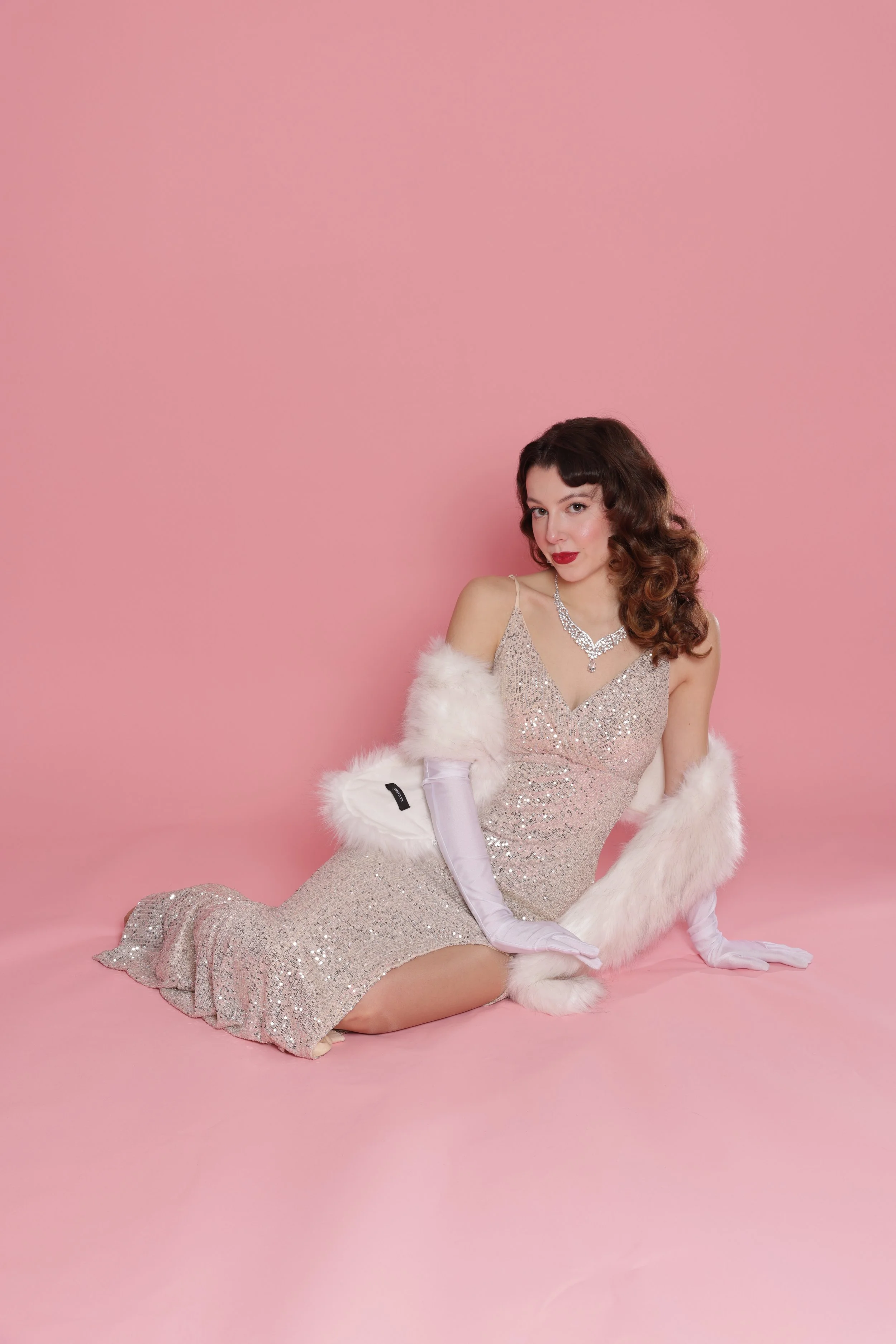 A woman in a glittering pink gown with a slit, wearing long white gloves, a white fur stole, and silver jewelry, sitting on a pink backdrop.