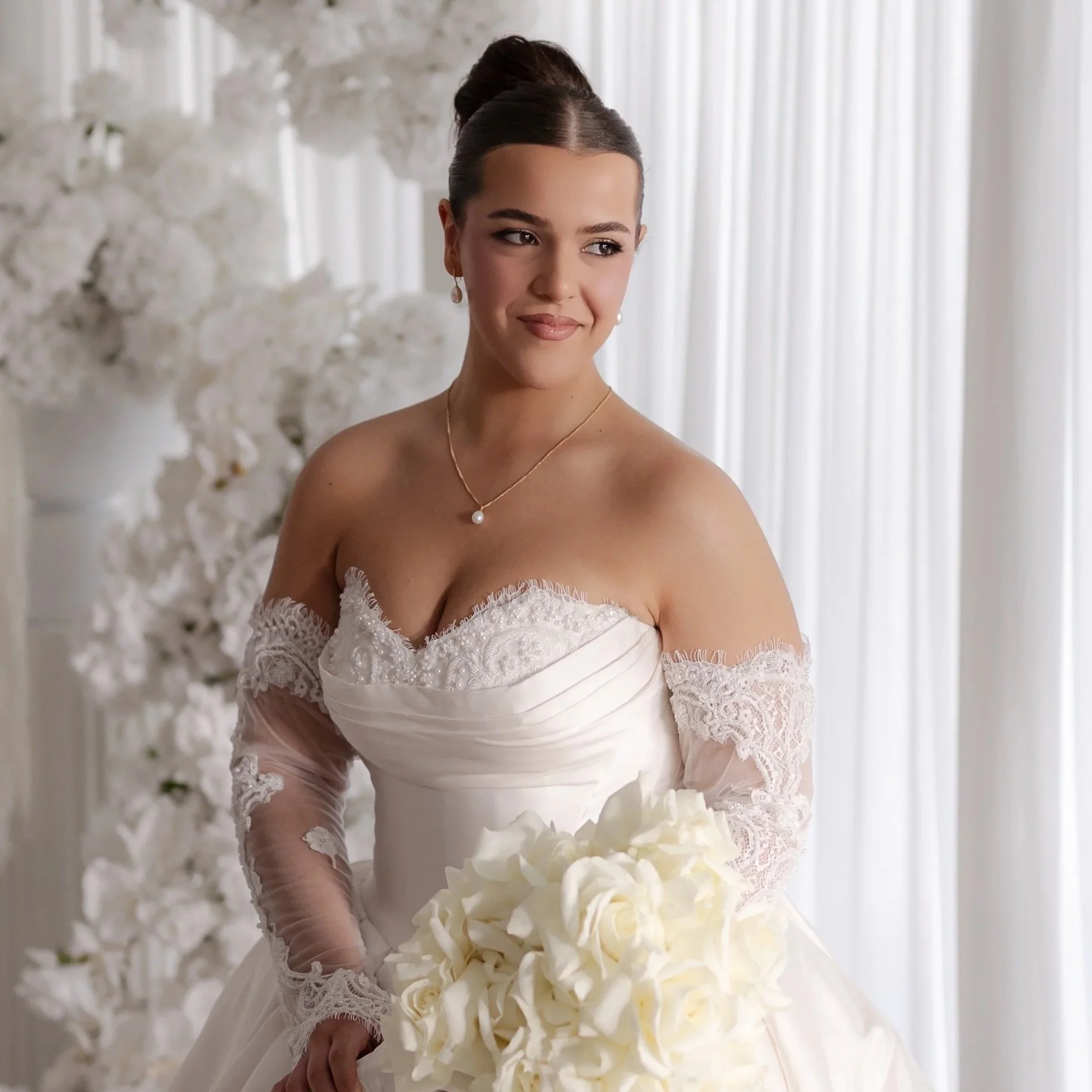 A bride in a white wedding gown holding a bouquet of white roses, standing in a bright room with white curtains and floral decorations.