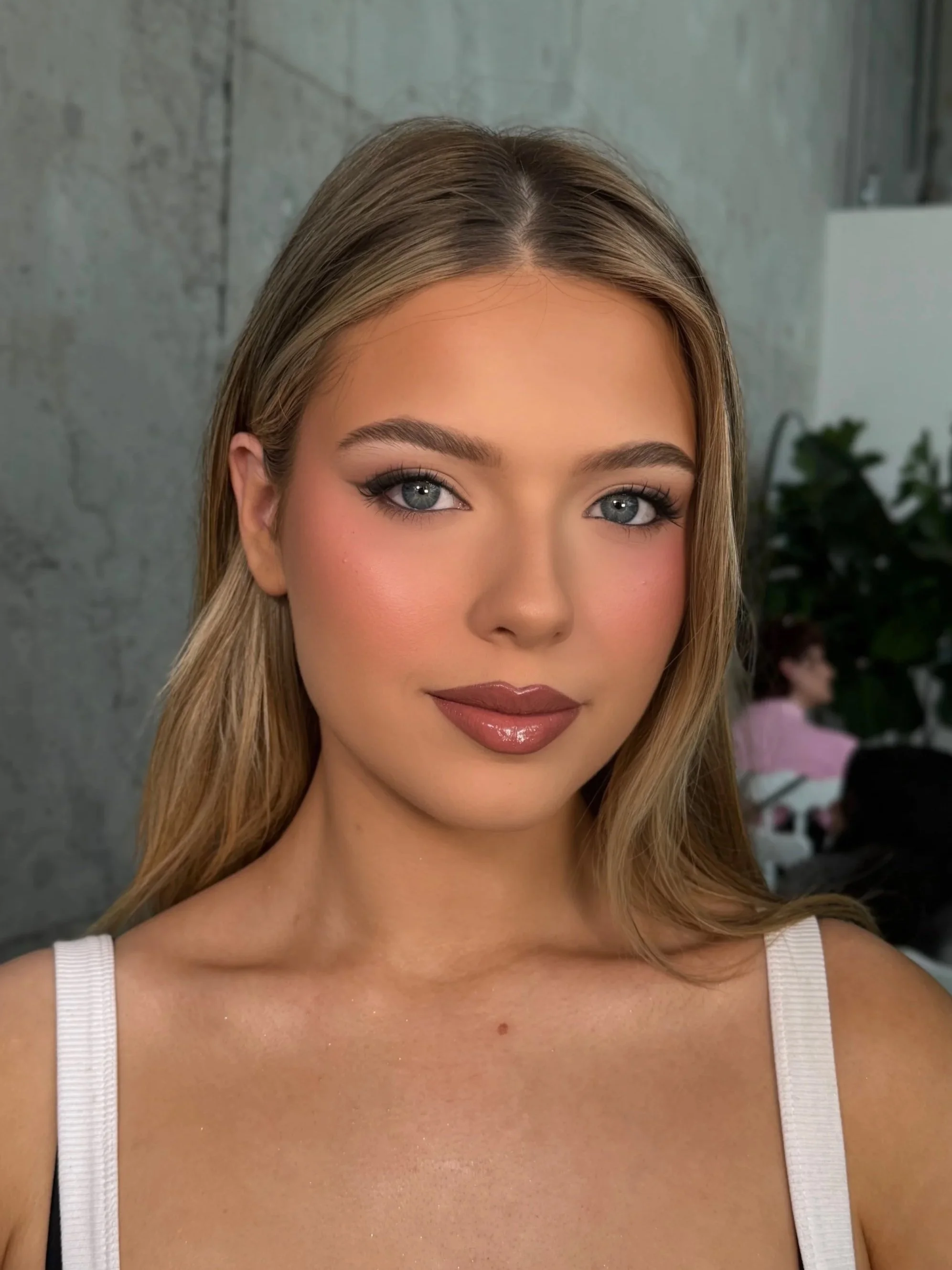Close-up of a young woman with blonde hair and blue eyes, wearing makeup with a natural background.