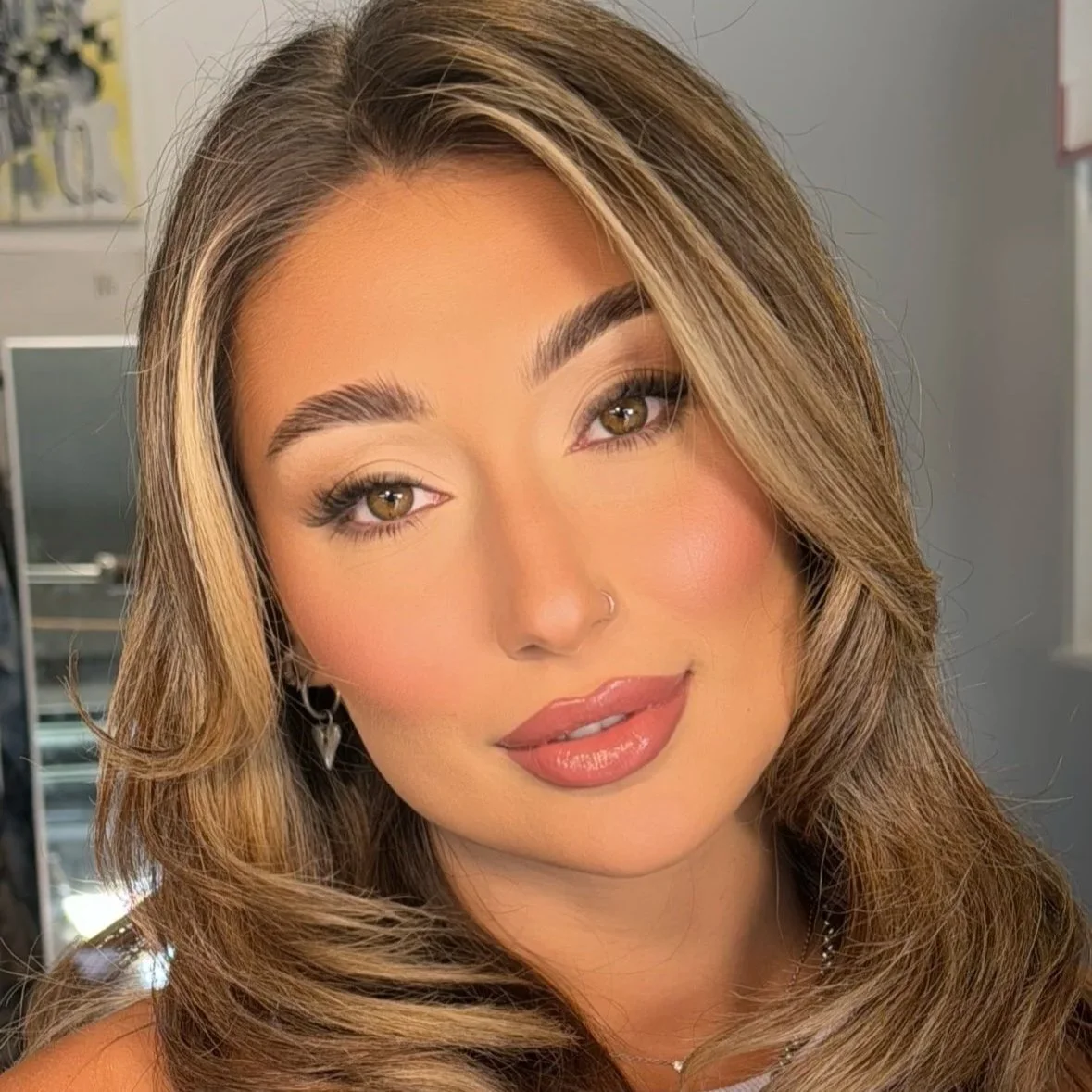 Close-up portrait of a woman with long, wavy light brown hair, wearing makeup with neutral tones, glossy lips, a nose ring, and earrings, in an indoor setting.