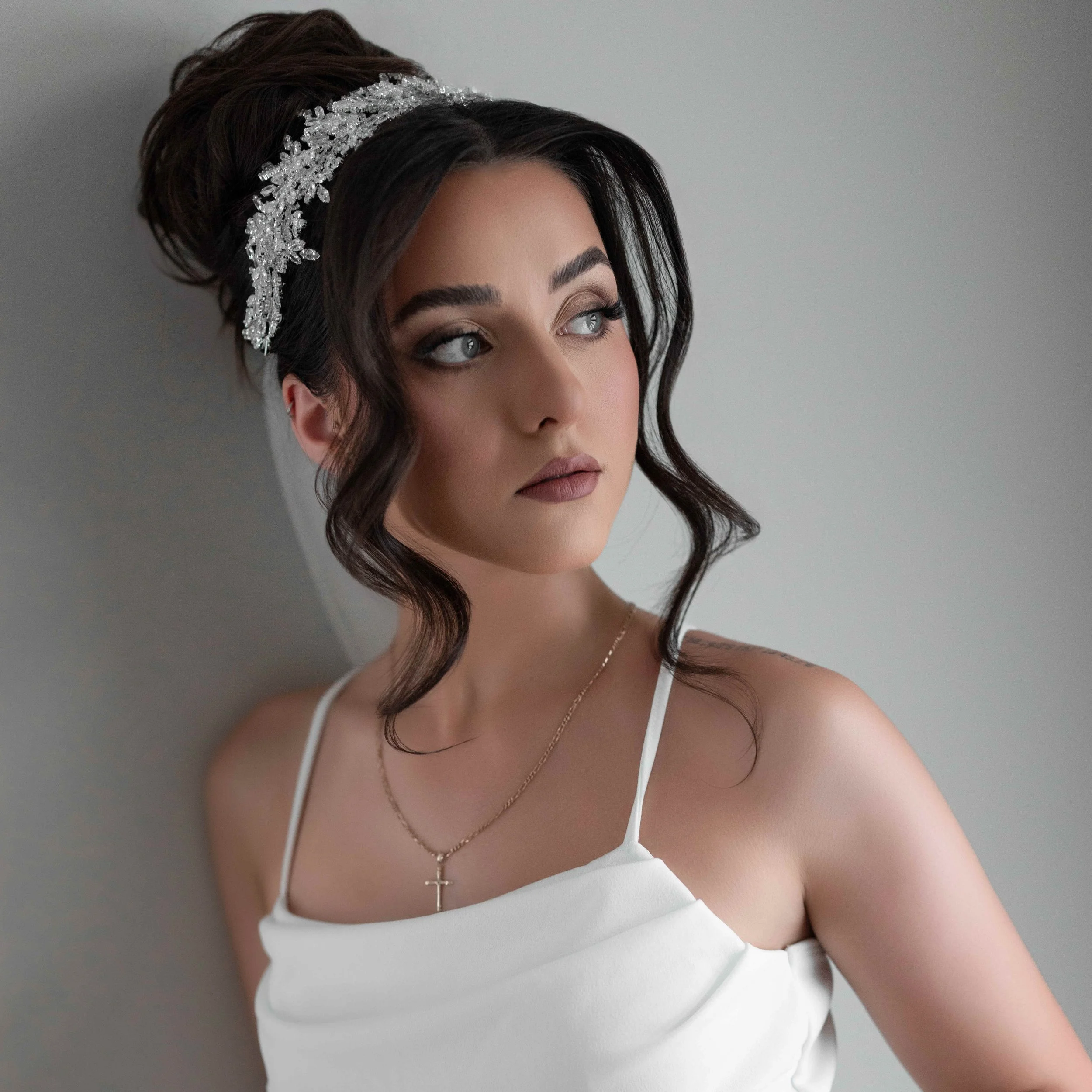 A woman with dark wavy hair wearing a white dress with thin straps, a silver tiara, and a gold cross necklace, looking off to her right with a serious expression.