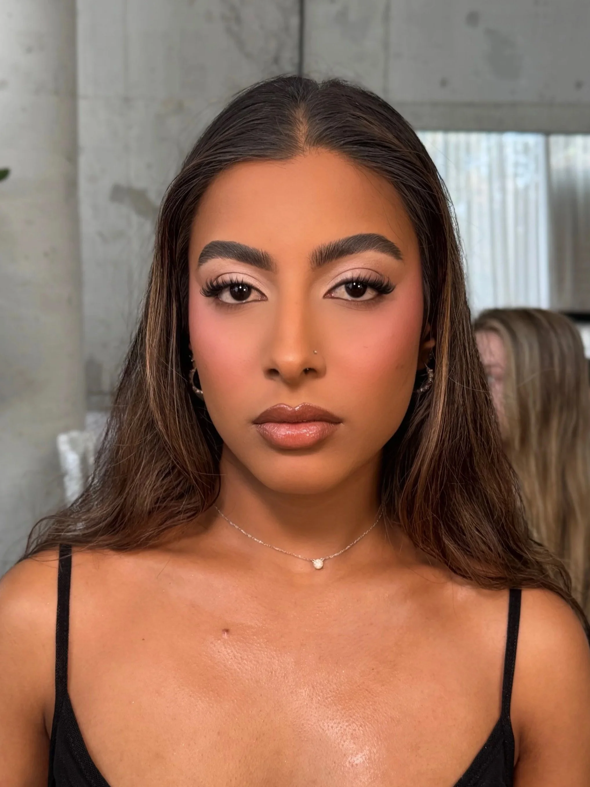 Close-up of a young woman with long brown hair, wearing makeup, a nose piercing, earrings, a delicate necklace, and a black spaghetti strap top, standing indoors with a modern background.