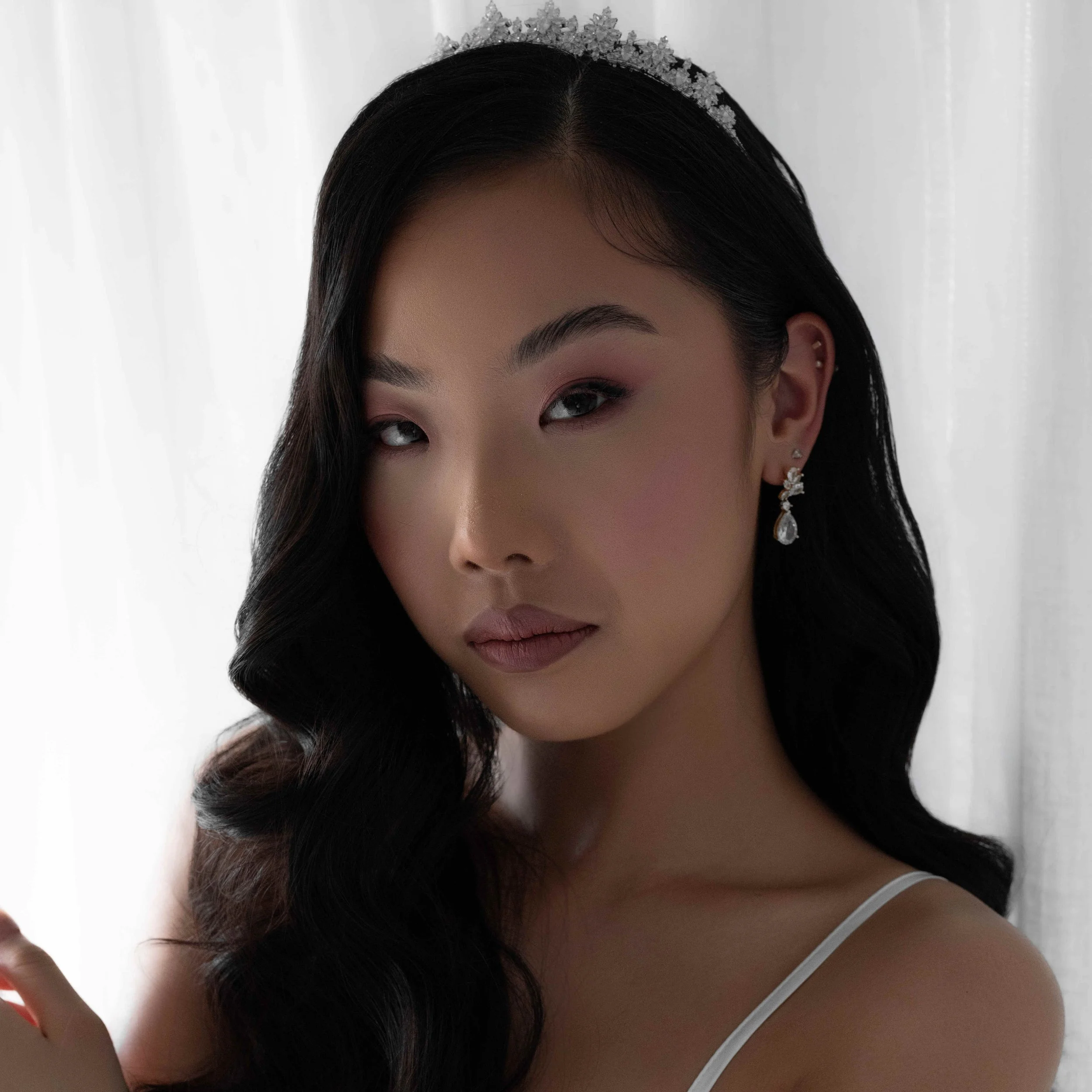 A woman with long, wavy black hair wearing a tiara and elegant earrings, with makeup and a white strap dress, standing in front of a white curtain.