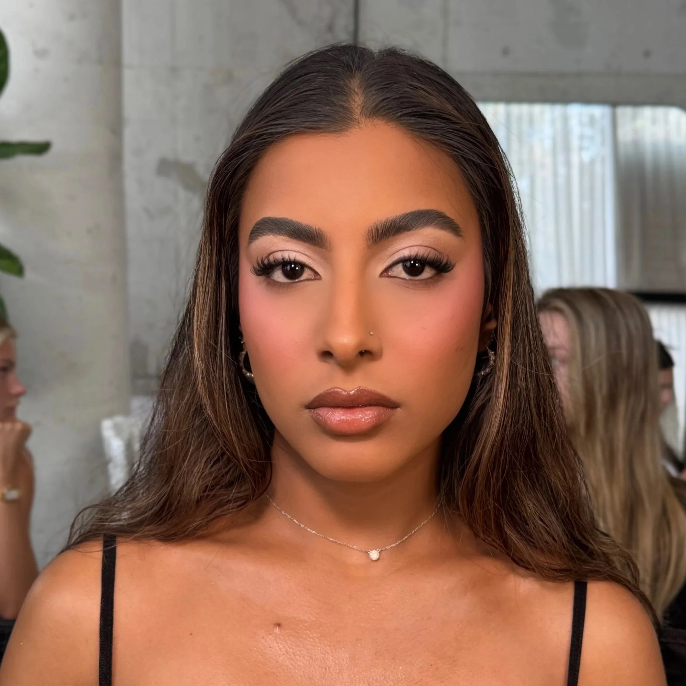 A young woman with long, dark hair, wearing makeup and a black spaghetti strap top, stands indoors in front of a mirror with other people in the background.