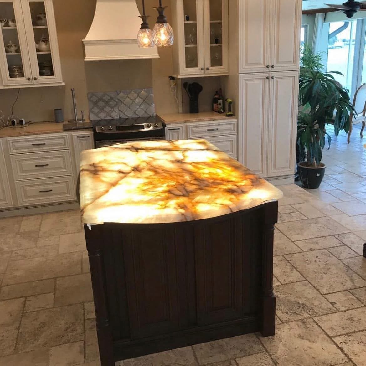 Kitchen with cream-colored cabinets, a black stove, and a lit pendant light fixture. A kitchen island with a glowing onyx or marble top is in the foreground. There is a large potted plant near a glass door or window at the far right.