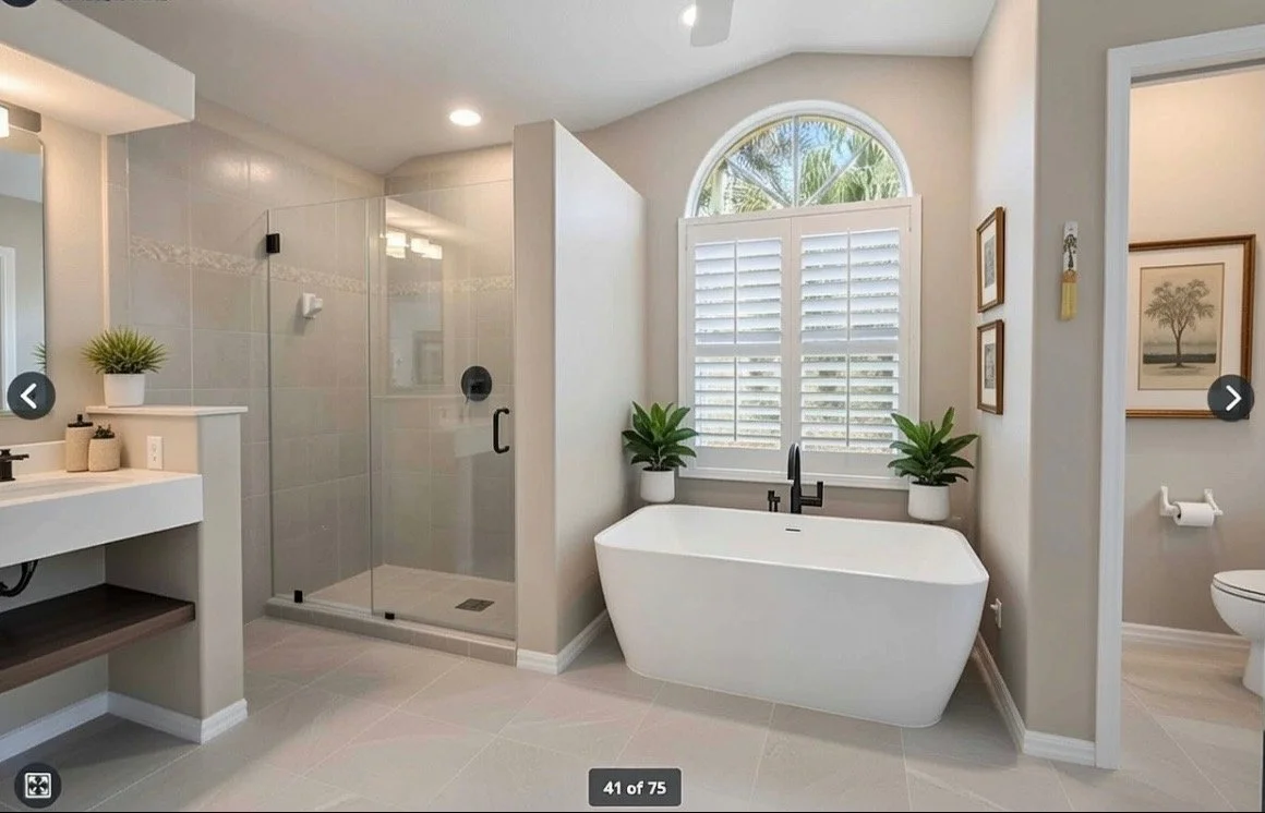 Modern bathroom with a glass-enclosed shower, a freestanding bathtub beneath a large window with white shutters, potted plants on the window sill, and a toilet with artwork on the wall.