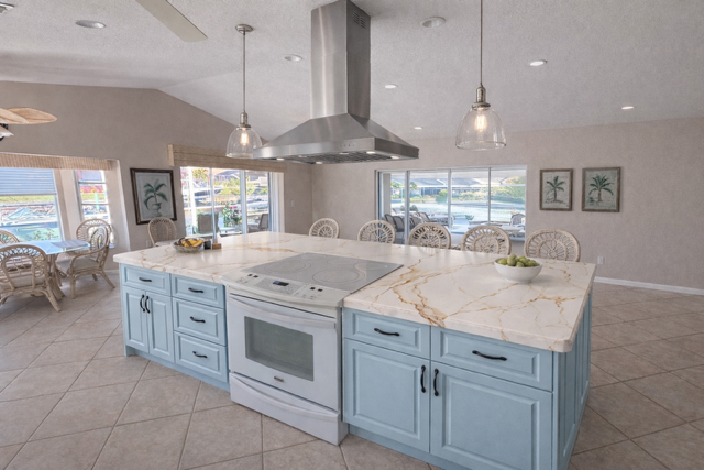 Modern kitchen with a large island featuring a white marble countertop, light blue cabinets, a stainless steel range hood, an electric stove, and pendant lighting. Large windows provide natural light and a view of parked cars outside.