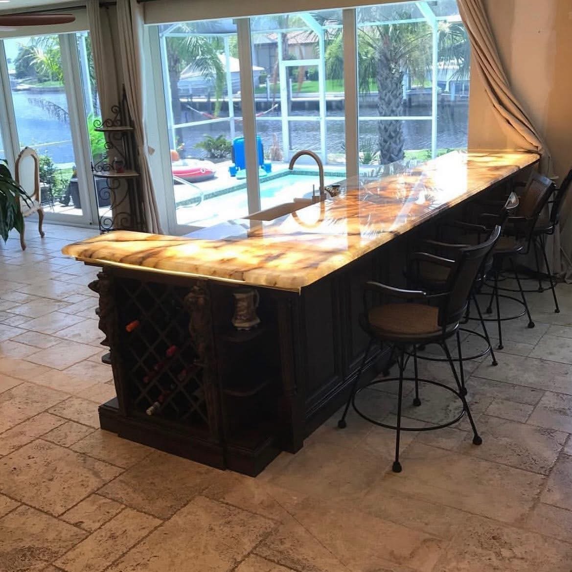 Kitchen island with a colorful, glossy marble countertop, black cabinetry, and bar stools in front of large windows overlooking a backyard with a pool, palm trees, and outdoor furniture.