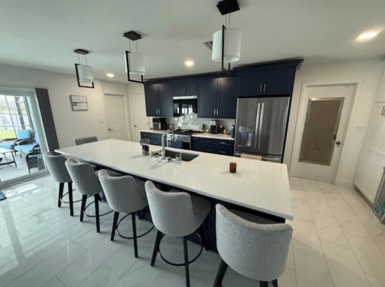 Modern kitchen with white island counter, bar stools, dark blue cabinets, stainless steel refrigerator, and pendant lighting.