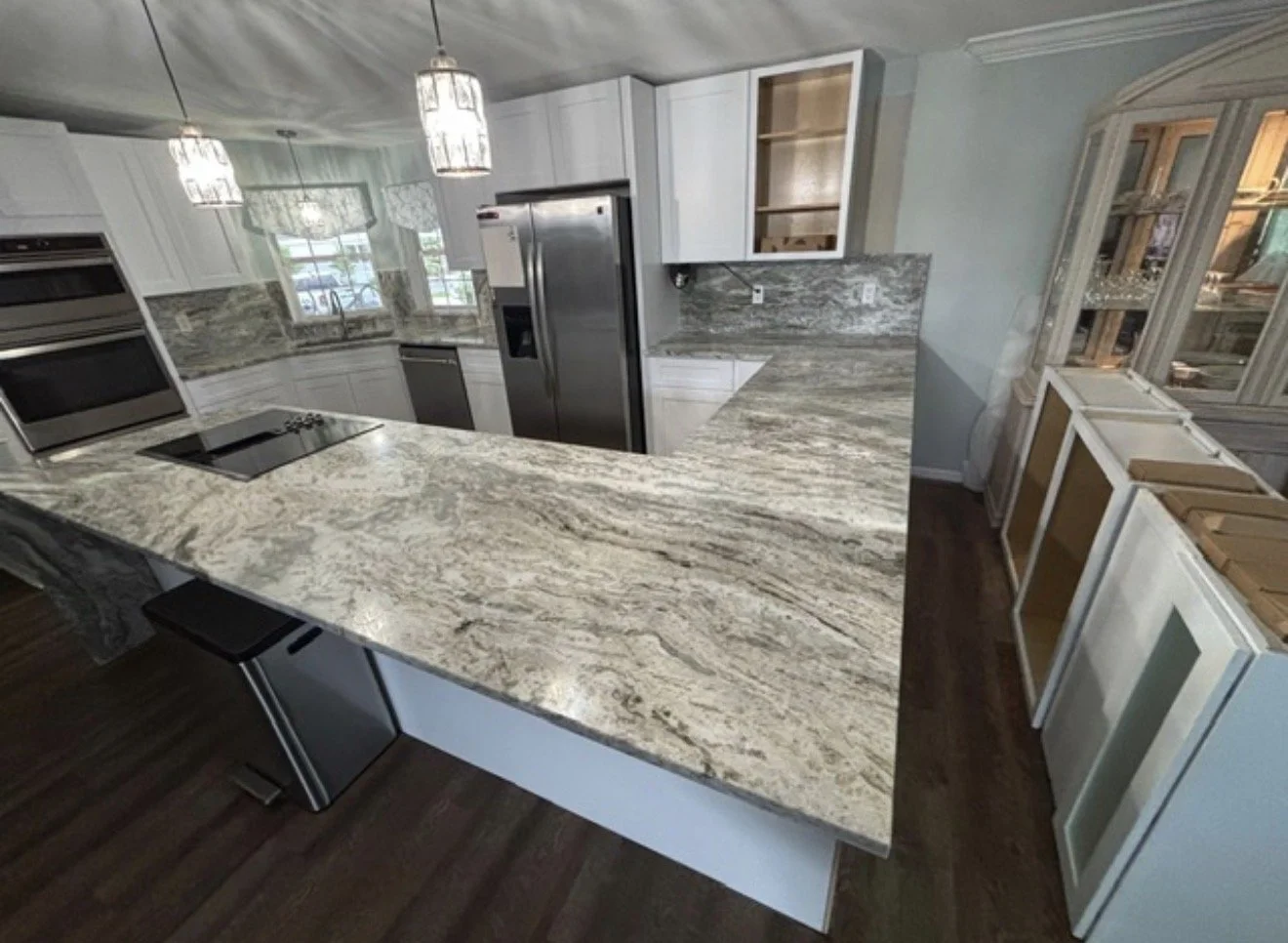 Kitchen with marble countertops, stainless steel appliances, white cabinets, and hardwood flooring. There is a kitchen island and a built-in oven with a windowed cabinet above.