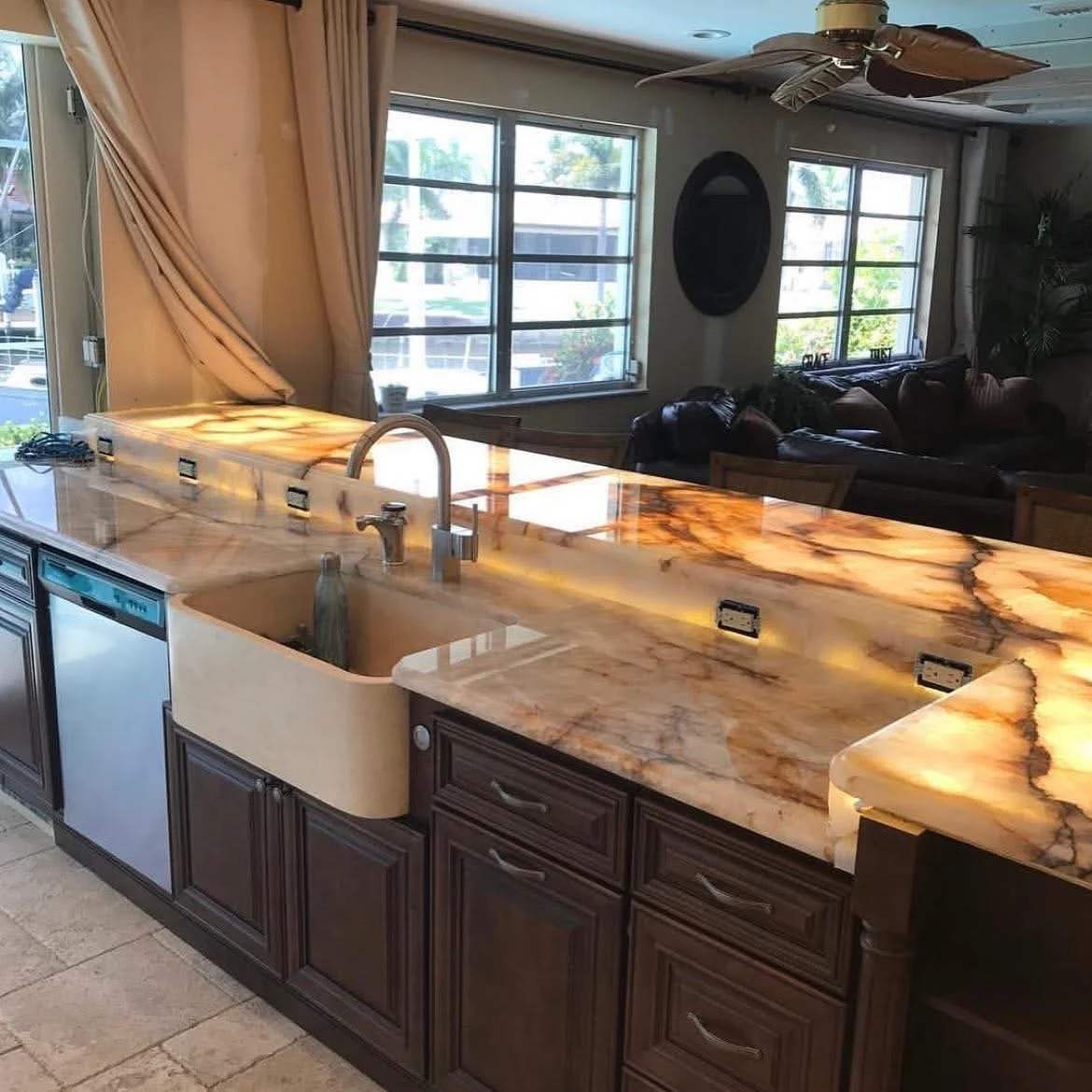 Kitchen with brown wooden cabinets, a onyx countertop with brown and cream patterns, a beige sink, and a modern faucet. In the background, living room with large windows, curtains, and a seating area with leather couches.