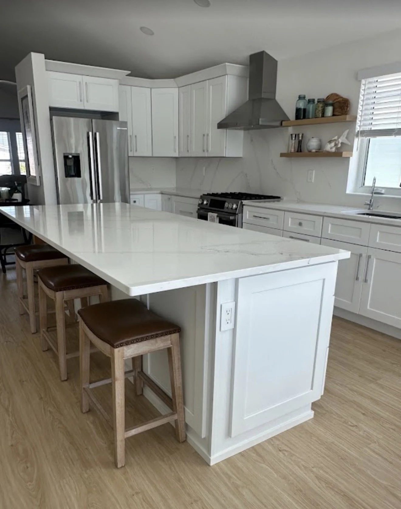 Modern white kitchen with marble island, stainless steel refrigerator, oven, and cooling range, open shelving with jars, window with blinds, and light hardwood flooring.