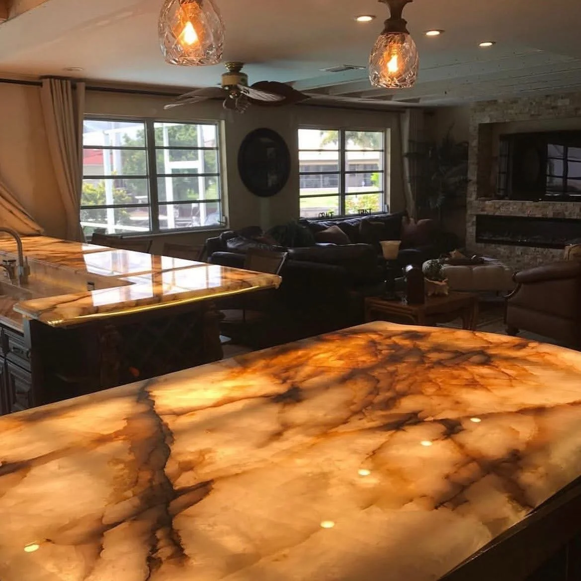 Living room with orange and brown marbled countertops, dark leather sofas, large windows, a stone fireplace, and hanging pendant lights.