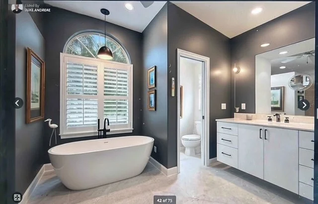 Bathroom with a freestanding bathtub near a window with plantation shutters, dark gray walls, and a white vanity with a large mirror, in a modern home.