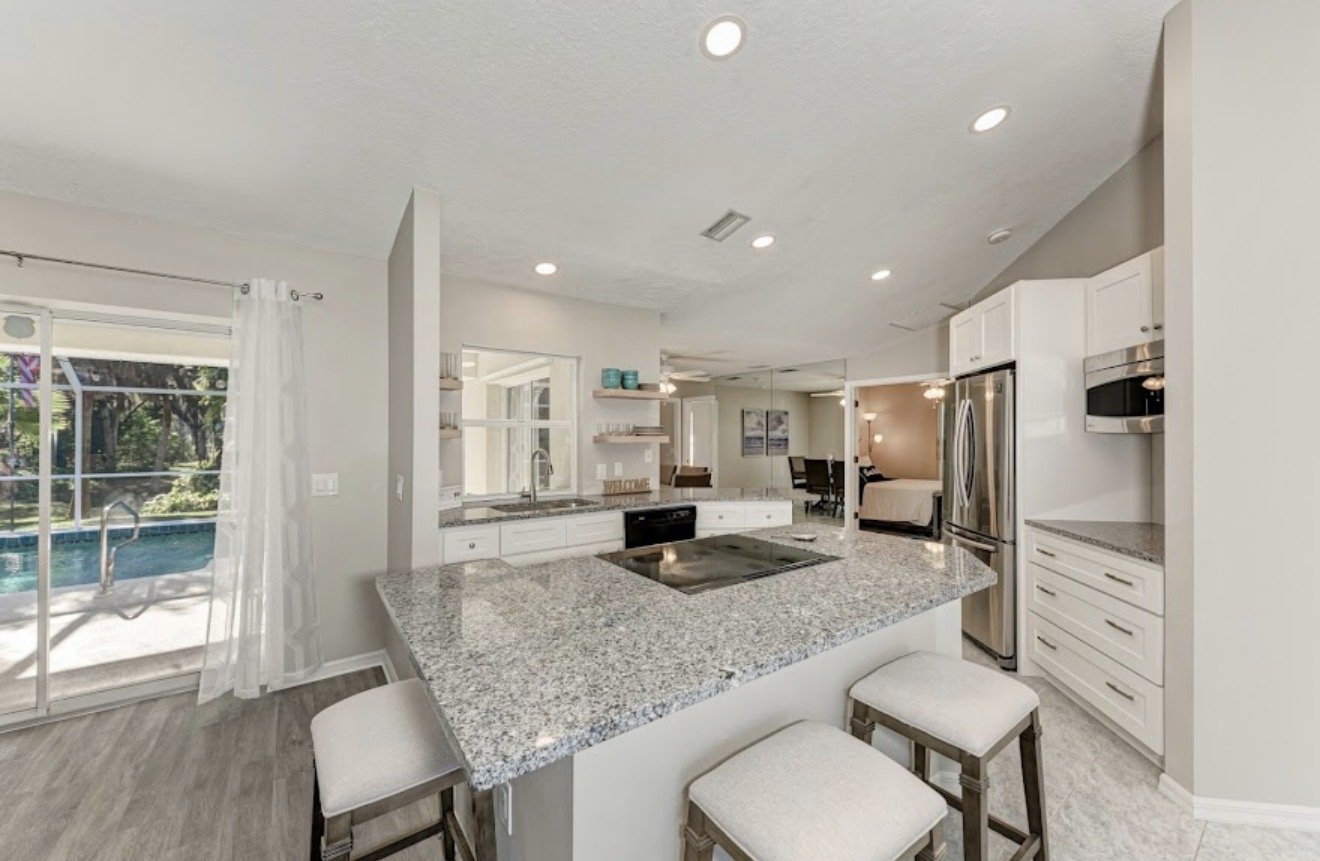 Modern kitchen with granite countertops, white cabinets, stainless steel appliances, an island with seating, and a view of a screened-in pool area outside.