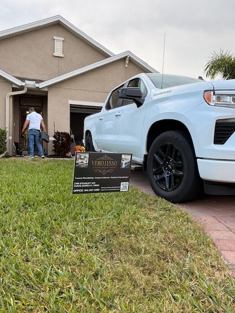 A white pickup truck parked on a driveway in front of a tan house. A person is standing near the house's front door. A sign in the grass next to the truck advertises Vero Lasso security and surveillance services.