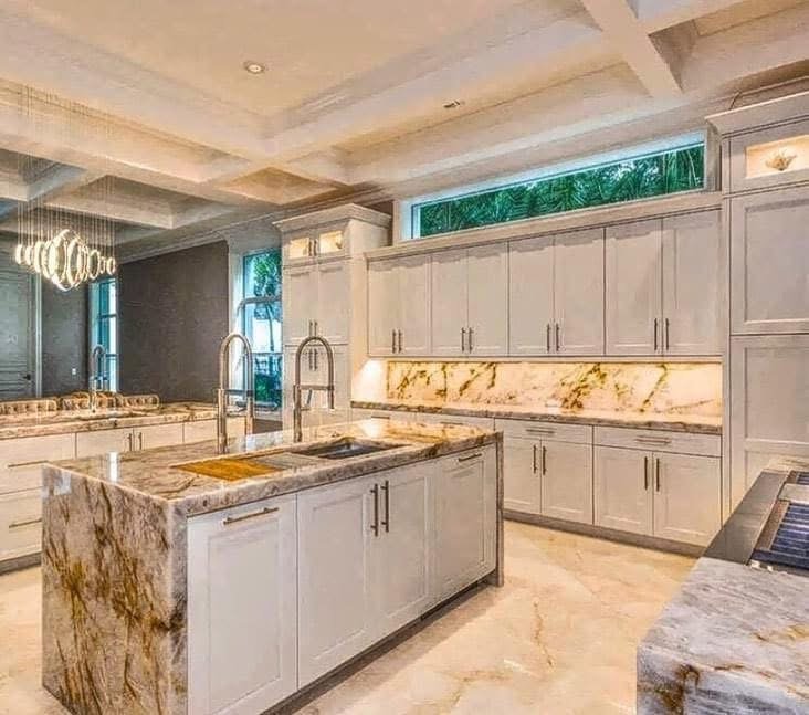 Modern kitchen with white cabinetry, marble countertops, and an island with a sink and faucet. Large windows and a decorative chandelier are visible.
