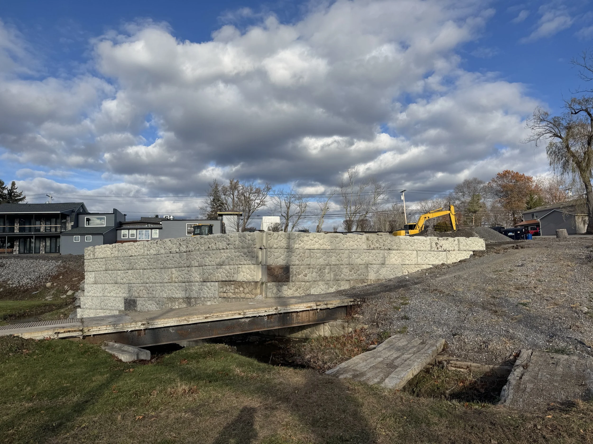Construction site with a yellow excavator on a mound of dirt behind a large stone retaining wall under cloudy sky.
