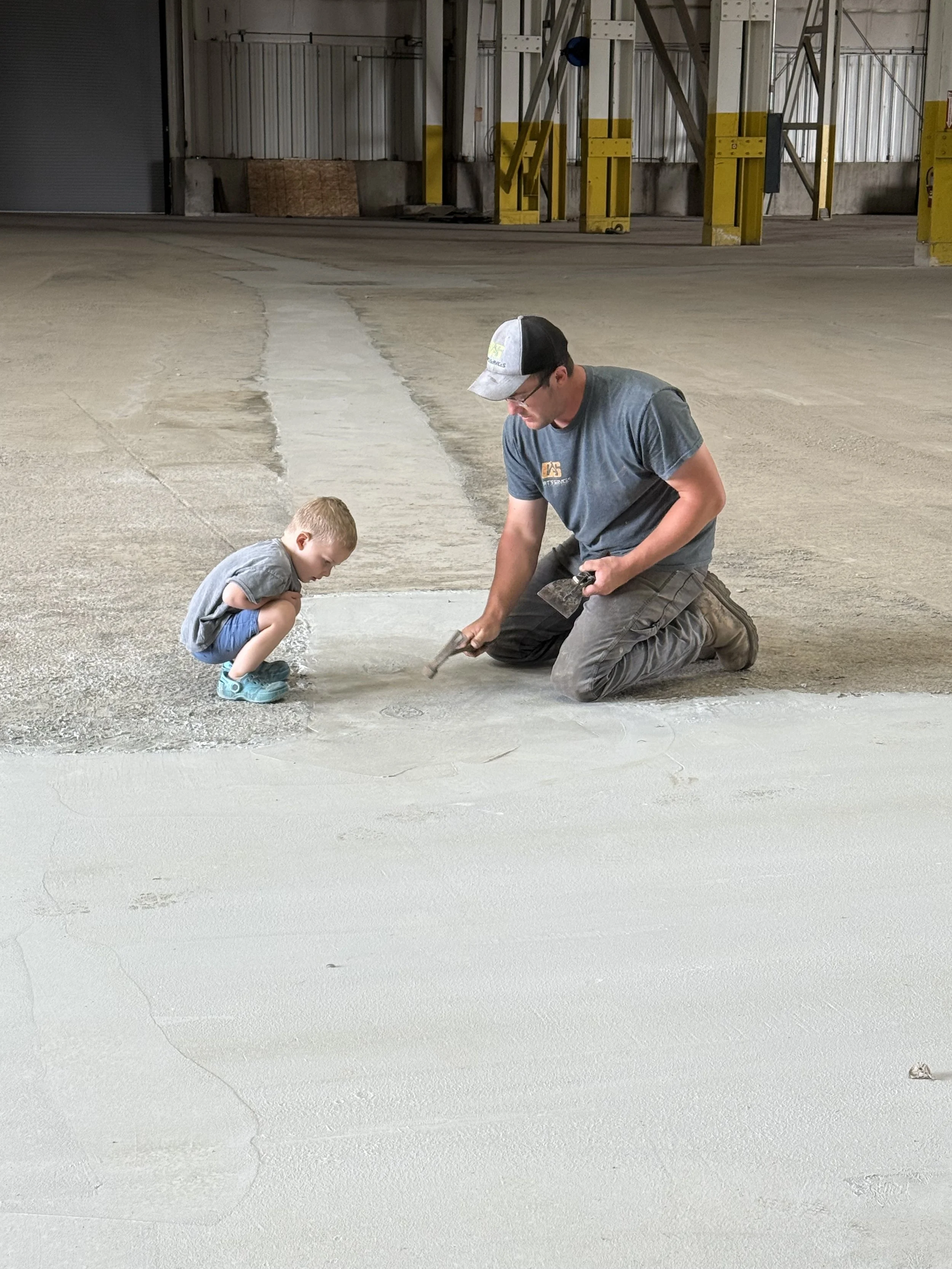 A man and a young boy working on concrete flooring inside a warehouse. The man is kneeling with a hammer, and the boy is squatting watching him.