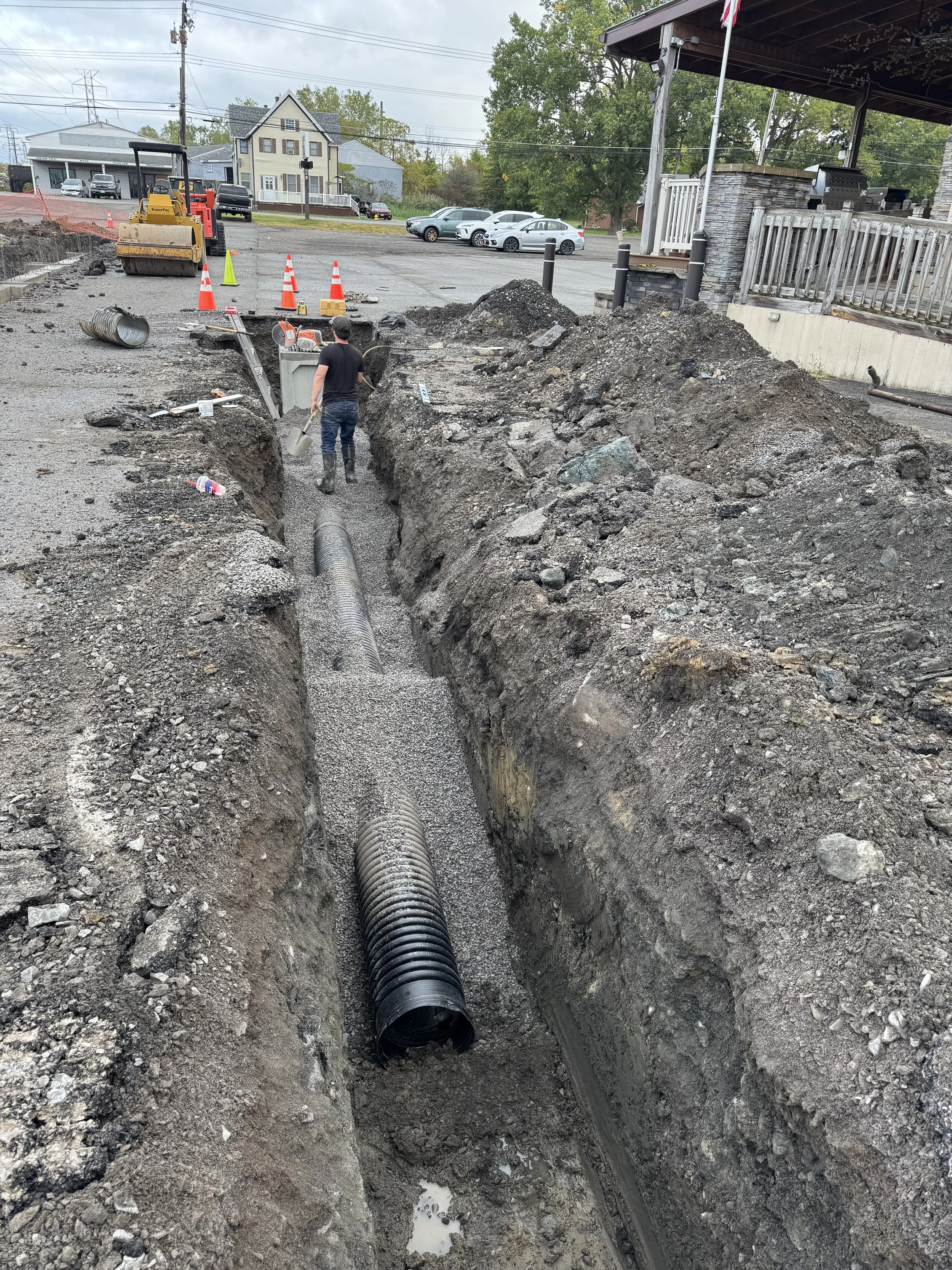 A worker in a black shirt and jeans is working in a trench on a construction site, installing underground pipes. Traffic cones are placed around the excavation area, with construction equipment and nearby residential and commercial buildings visible in the background.