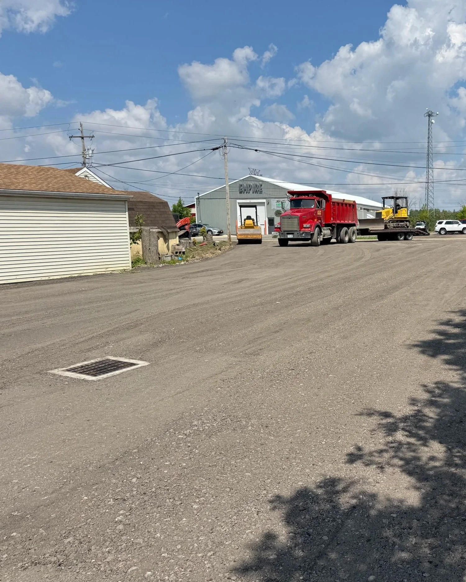 A dirt lot with construction vehicles, including a large red dump truck, a yellow bulldozer, and a roller, parked near a building with a sign that says 'EMPIRE,' under a partly cloudy sky.