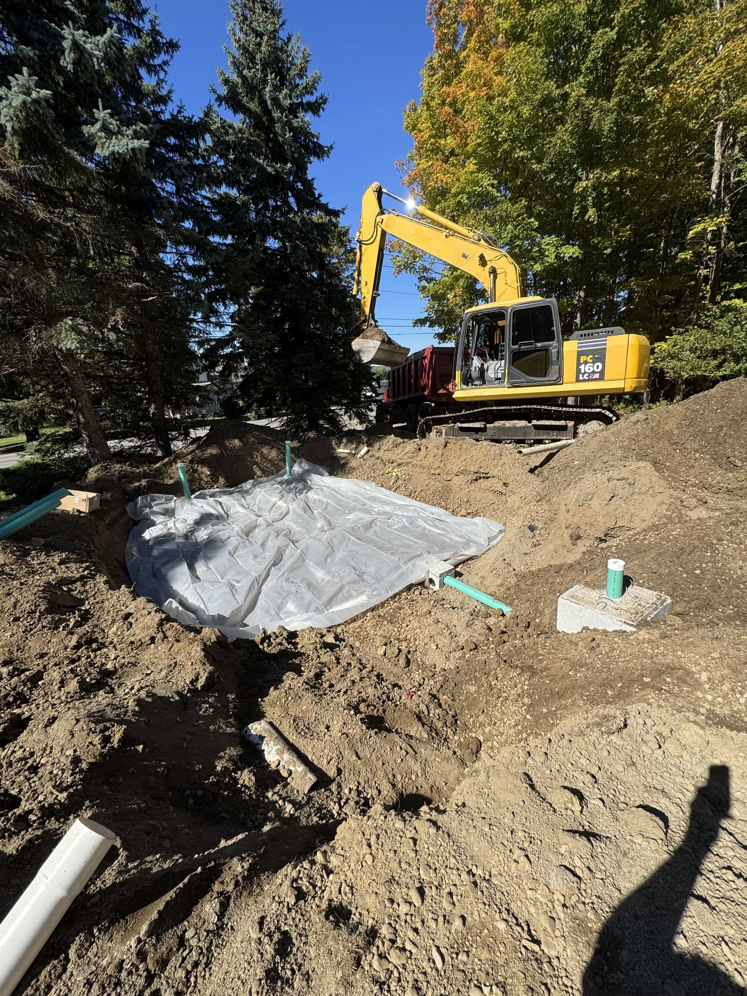 Construction site with a yellow excavator loading dirt into a red truck, surrounded by soil and pipes, with trees and blue sky in the background.