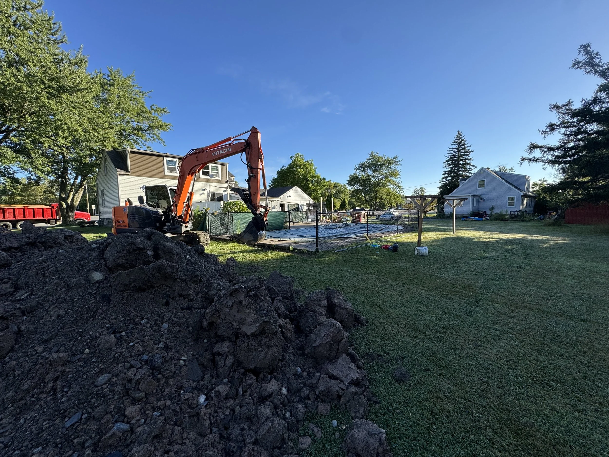 A backyard with a pile of dirt and an orange excavator on a sunny day.