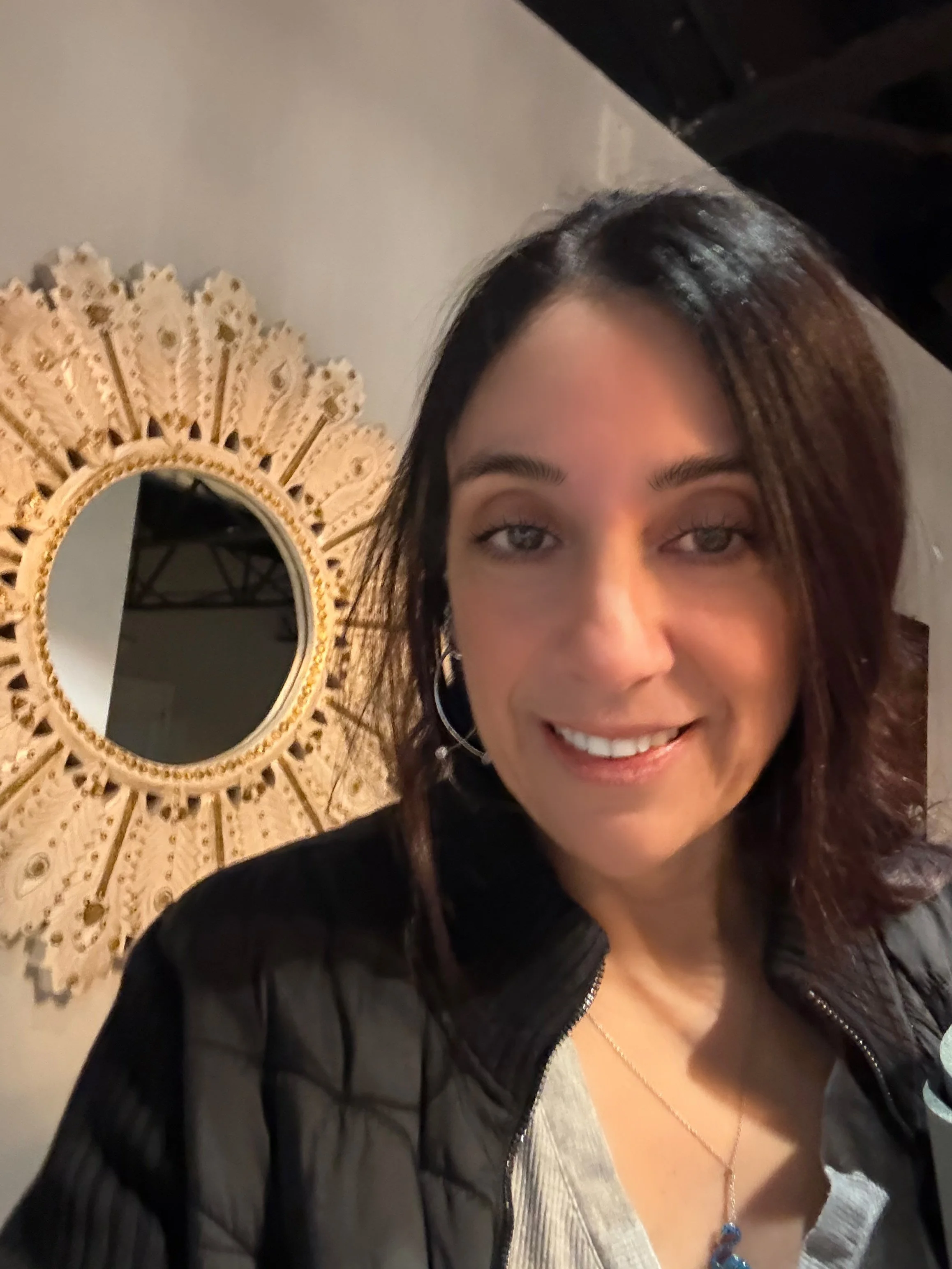 A woman with shoulder-length dark hair, light-colored eyes, and a smile. She is wearing hoop earrings, a necklace with a blue pendant, a light top, and a black jacket. Behind her is a decorative circular mirror with an ornate, sunburst design in gold.