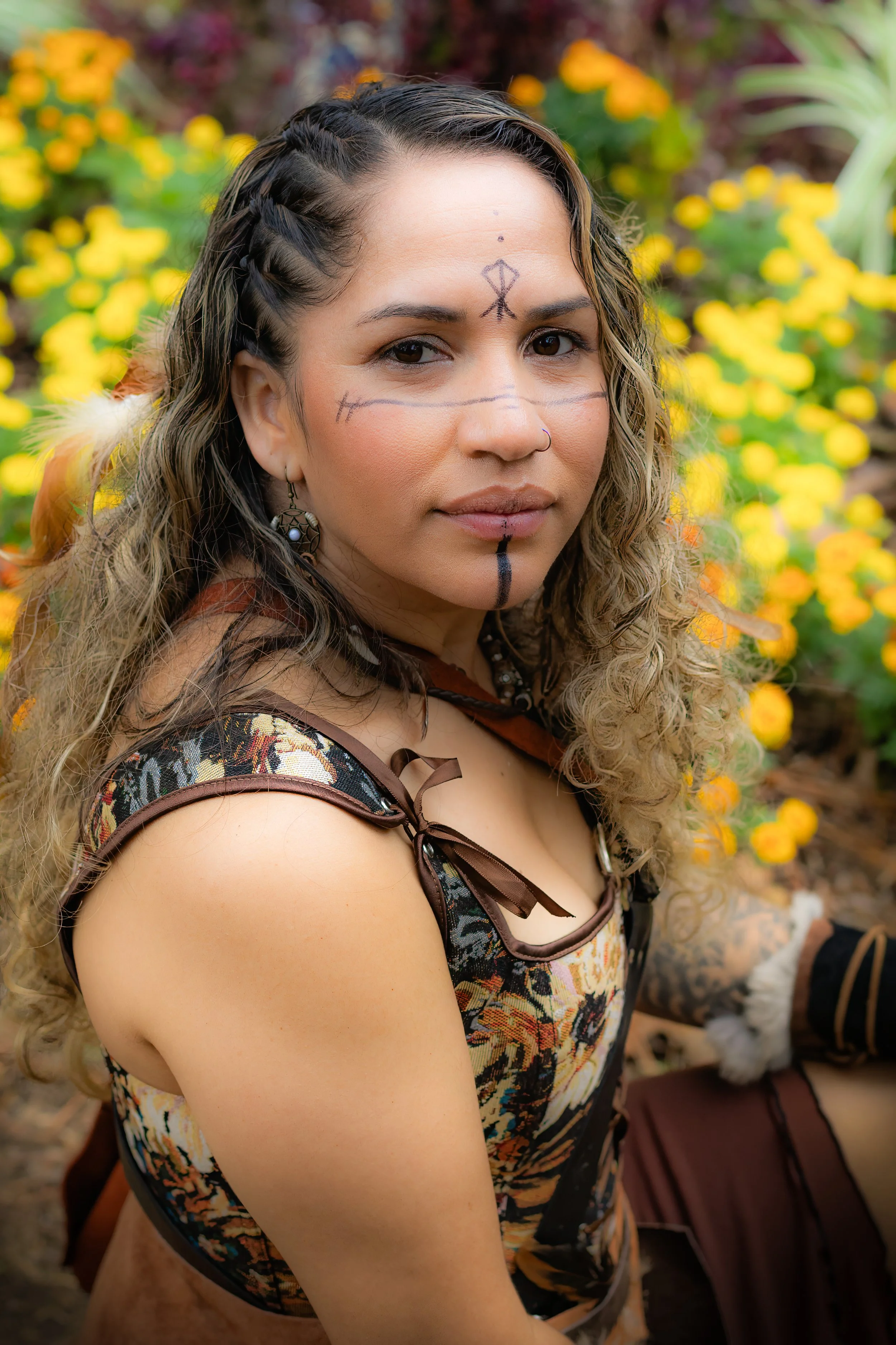 A woman with semi-long curly hair, tribal face paint, and traditional attire, sitting outdoors among yellow flowers.