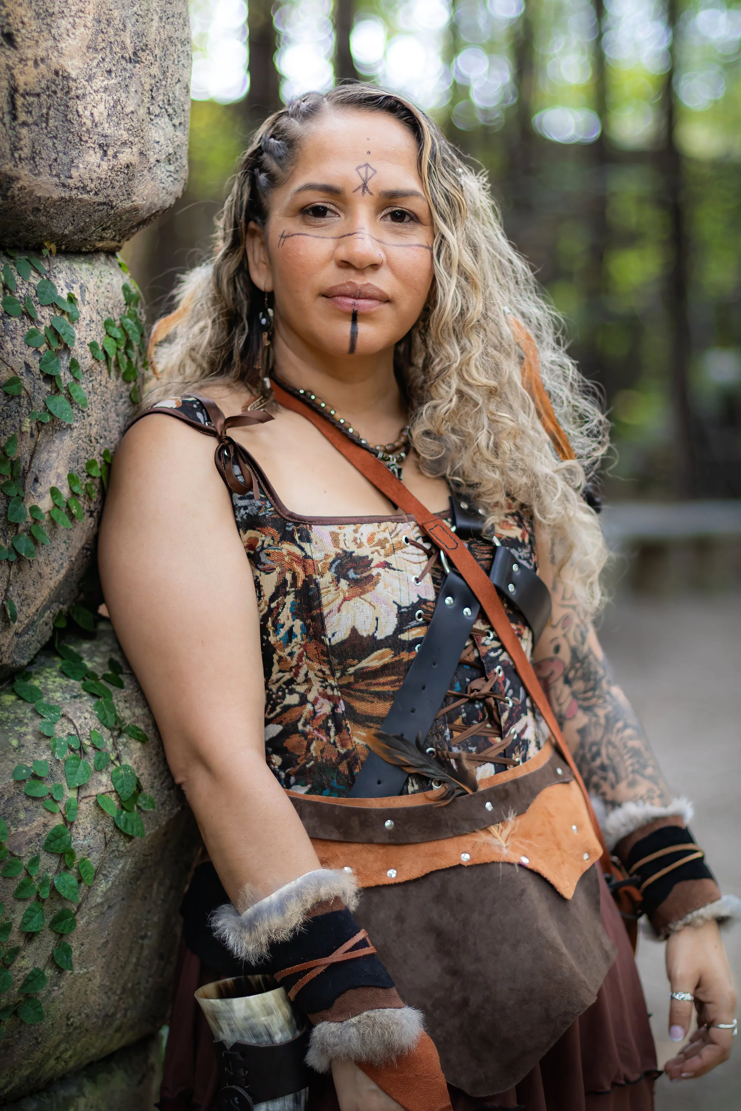 A woman in fantasy or tribal costume with face paint, standing outdoors with trees in the background, leaning against a stone structure.