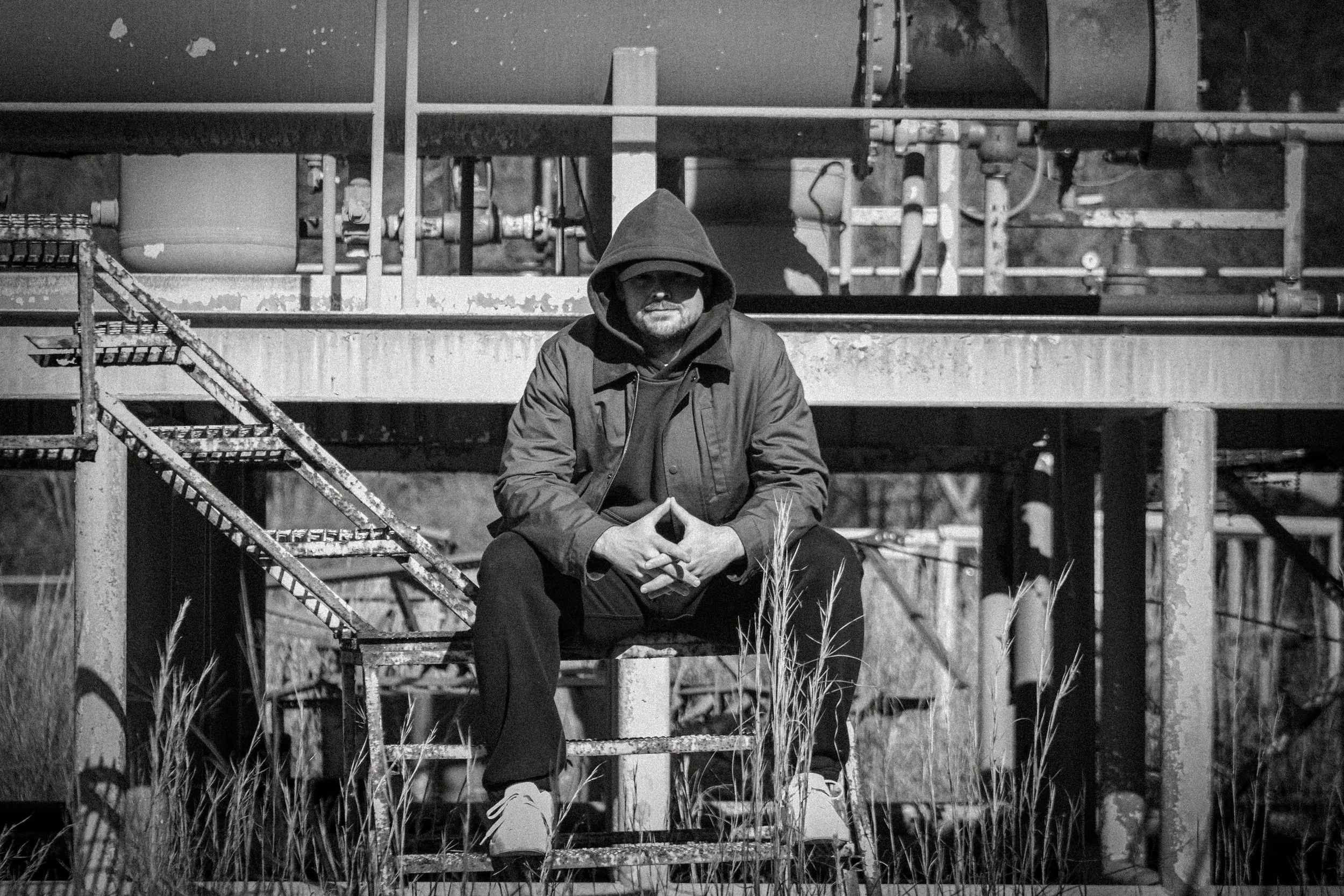 A man sitting on a rusty metal staircase in an industrial outdoor setting, wearing a hooded jacket and hoodie, with his hands clasped and fingers forming a triangle, surrounded by overgrown grass.