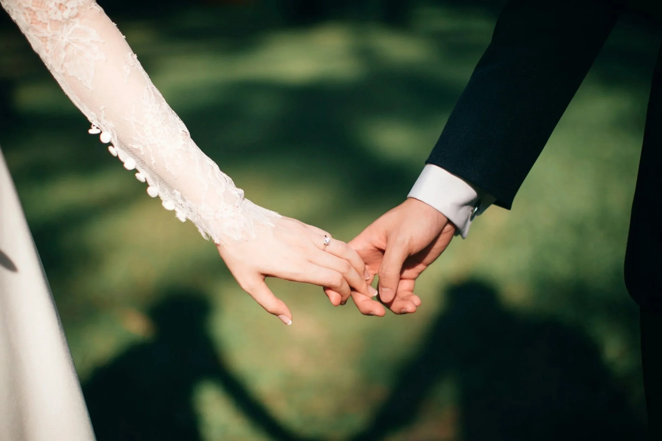 Close-up of a bride and groom holding hands outdoors, with the bride wearing a lace dress and the groom in a dark suit.
